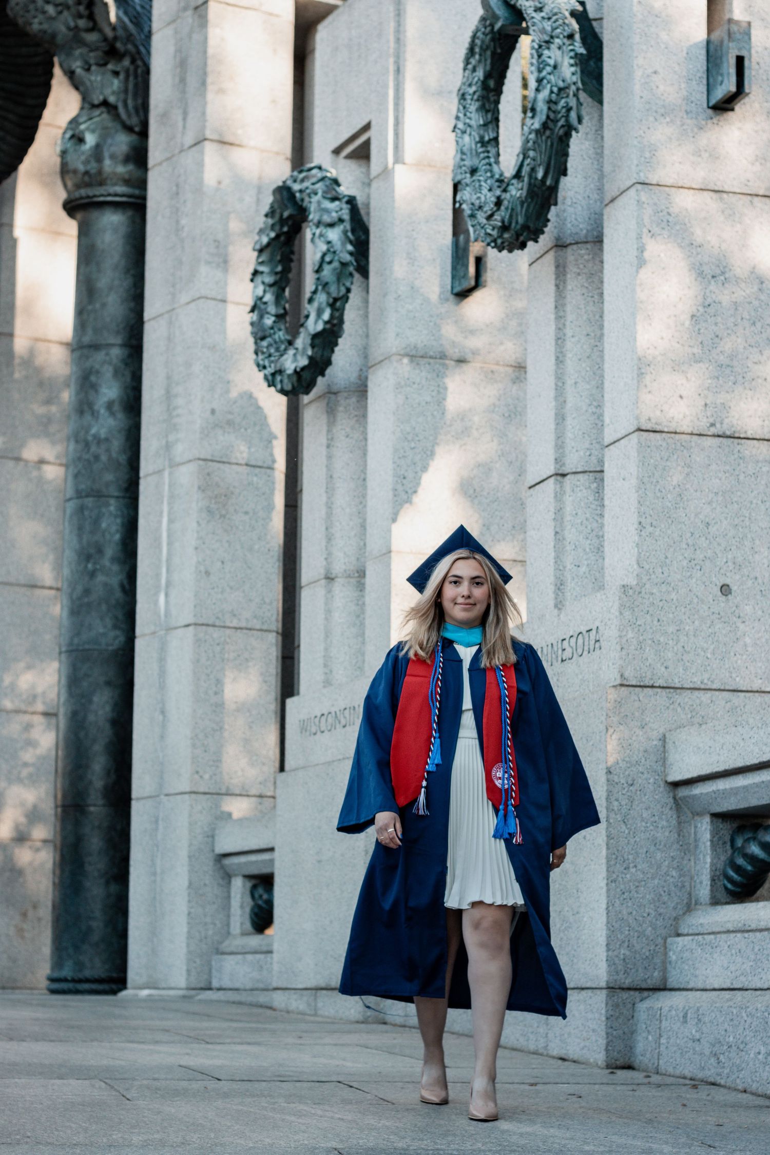 Woman in navy coat and red scarf poses among classical stone columns with warm sunlit architecture.