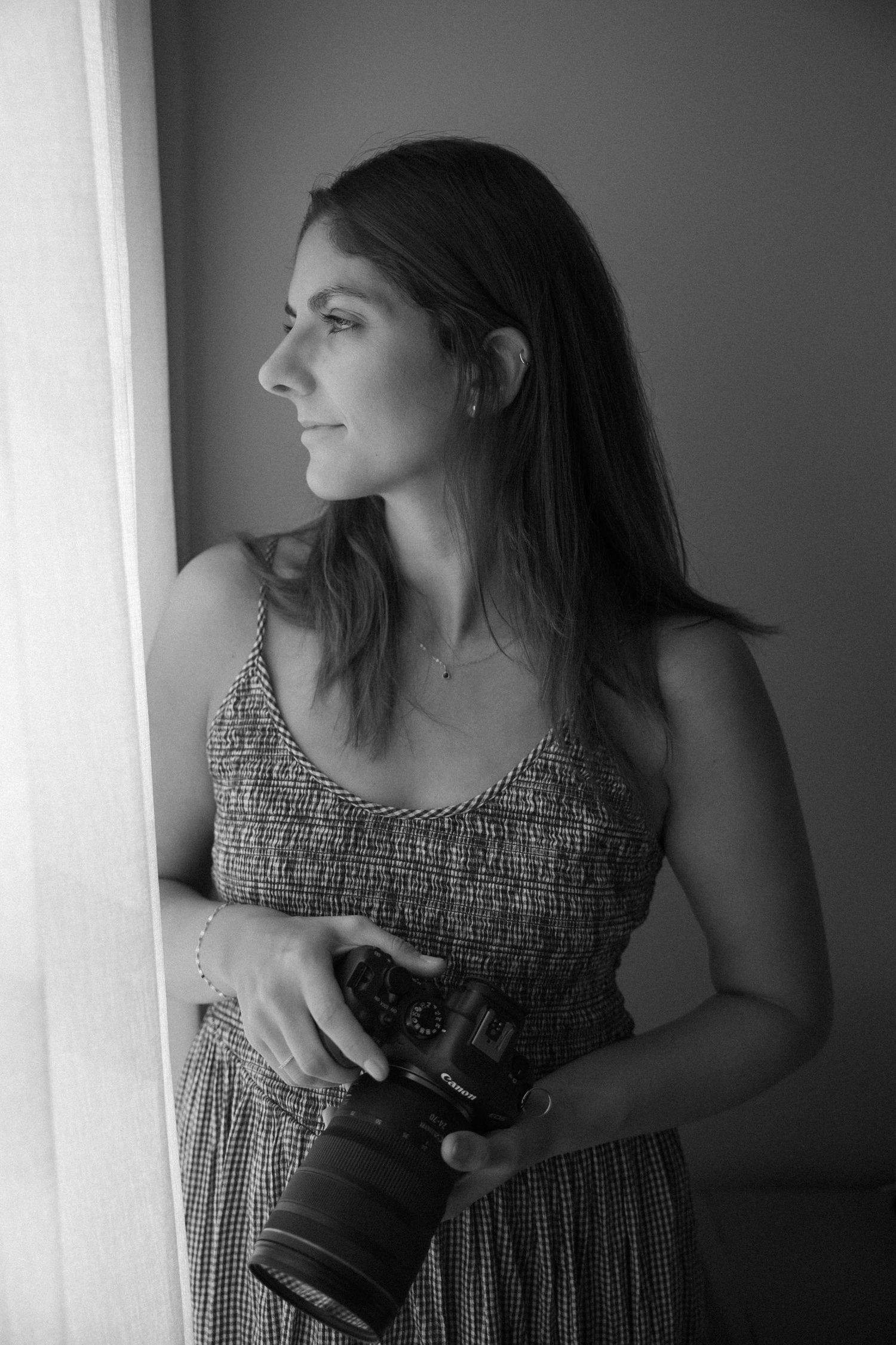 Black and white portrait of a woman holding a DSLR camera, wearing a sundress, gazing thoughtfully out a window.