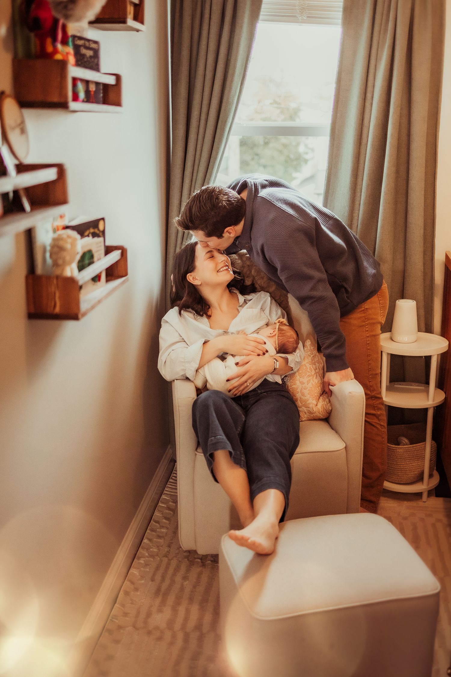A couple shares a romantic moment while sitting on a light-colored armchair in a cozy room with framed photos on the wall.