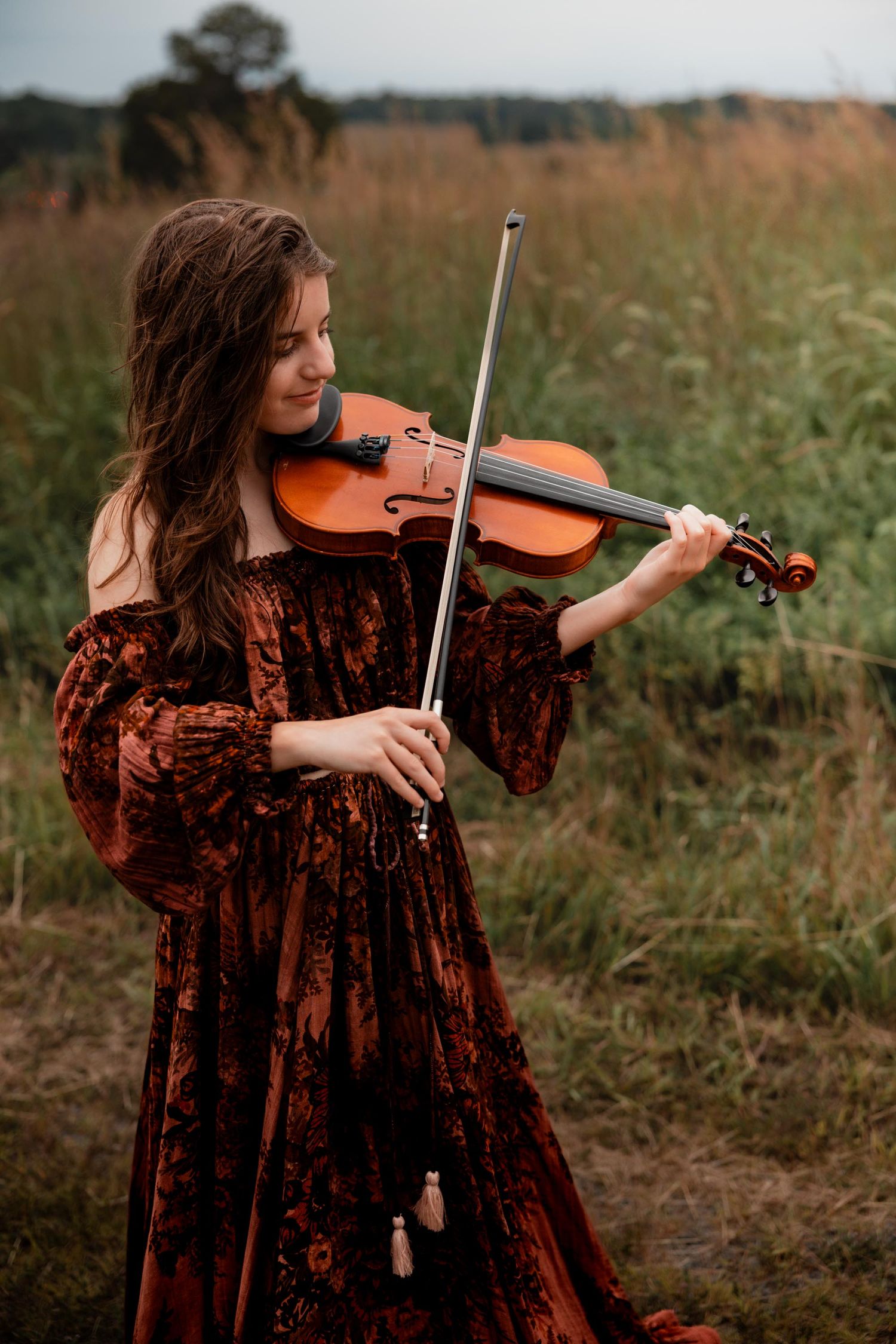 Young woman in a russet velvet off-shoulder gown playing violin in a lush green field.