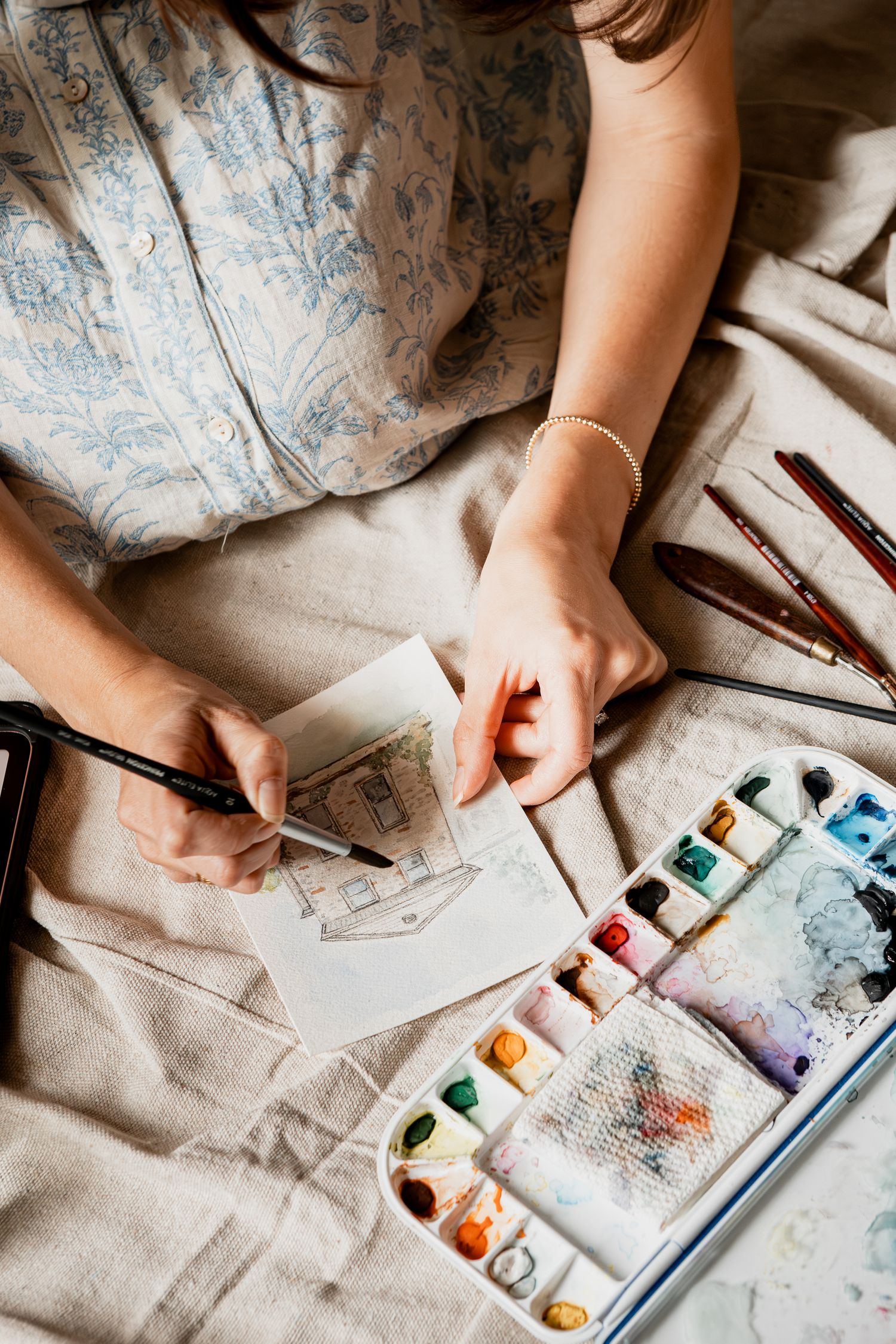 Artist painting with watercolors on white paper while sitting on a beige fabric surface with art supplies nearby.