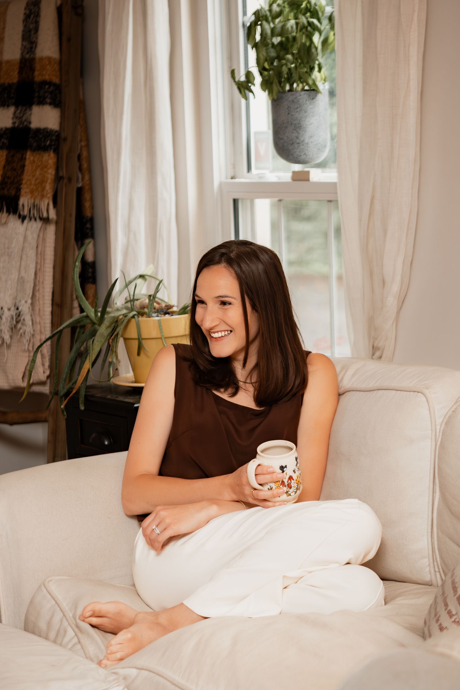 Person relaxes on white couch while enjoying coffee in bright sunlit room with hanging plants.