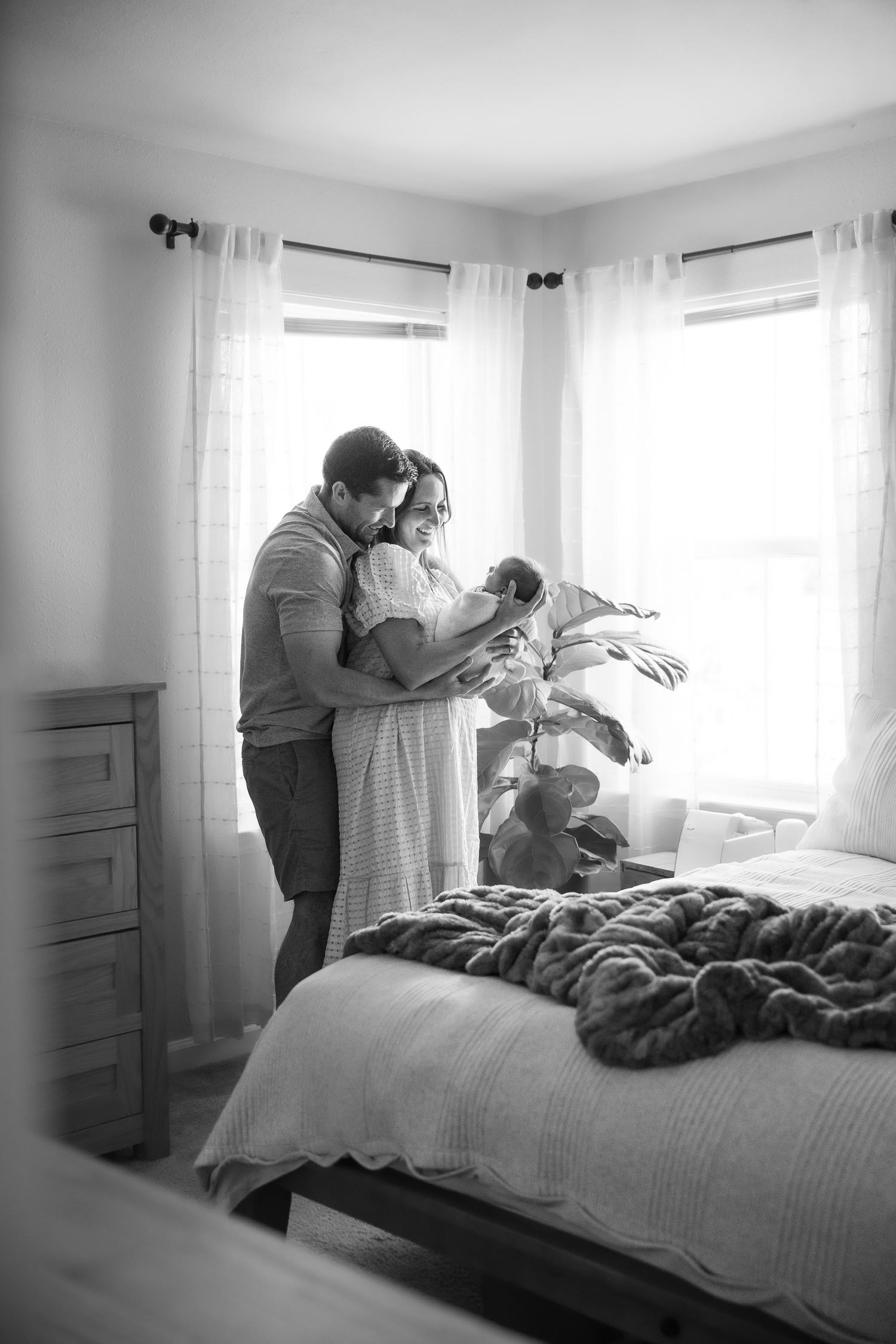Black and white photo of parents standing by bedroom window, lovingly gazing at their newborn baby.