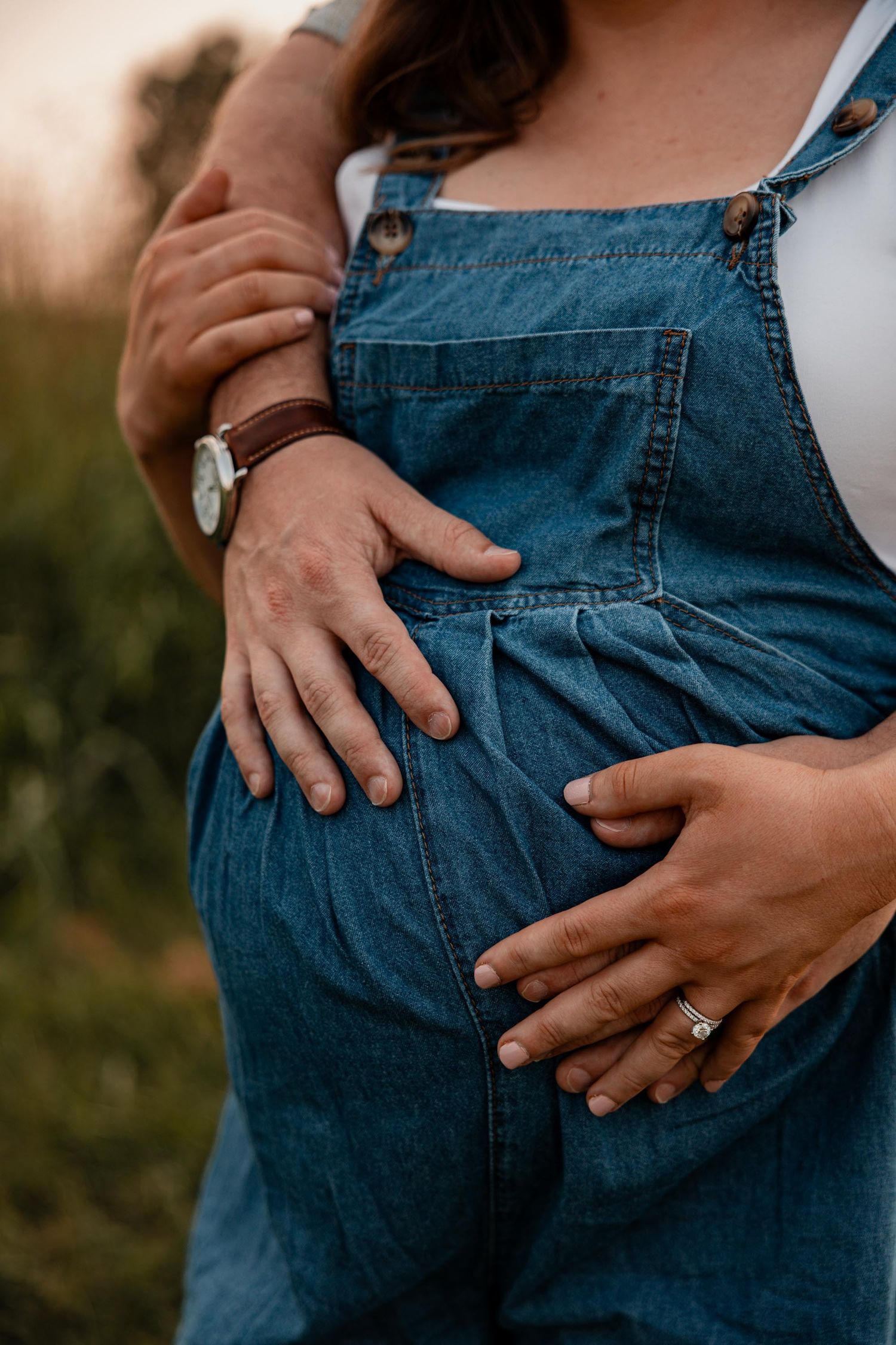 Pregnant woman in denim overalls cradling baby bump, partner embracing her from behind, both wearing rings, outdoor maternity photo.