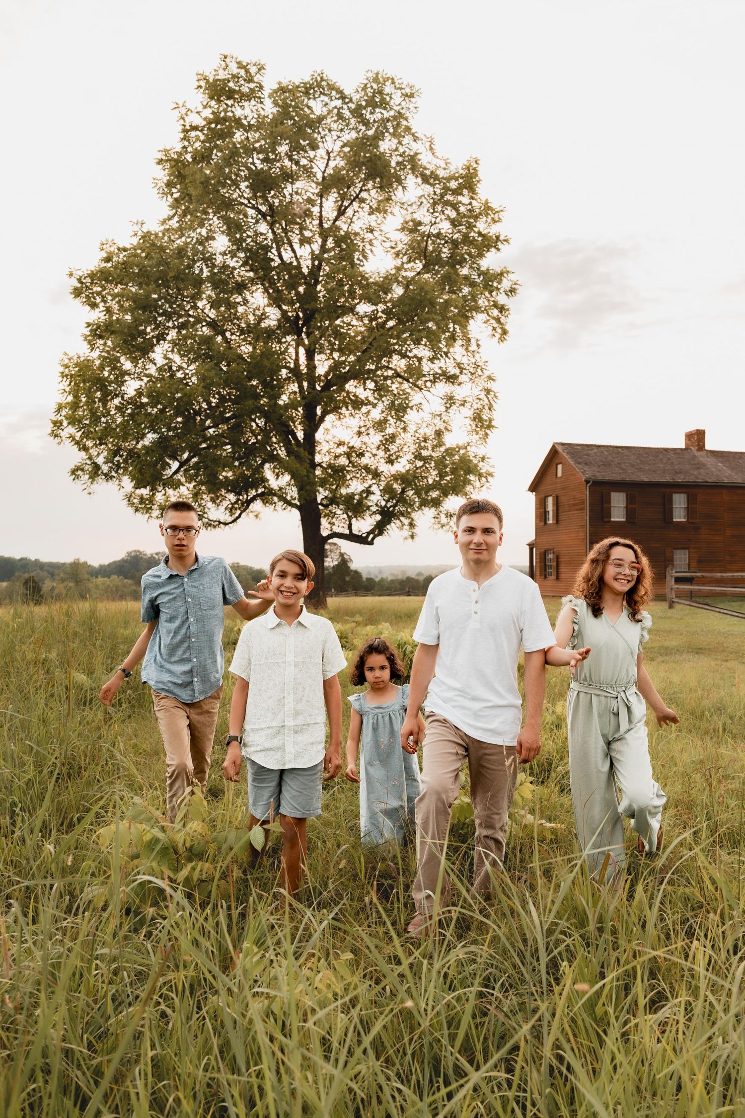 A blended family of five walks through a lush green field at golden hour near a rustic farmhouse.