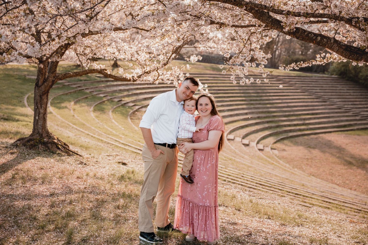 Family of three posing under blooming cherry blossom trees near an outdoor amphitheater in spring.