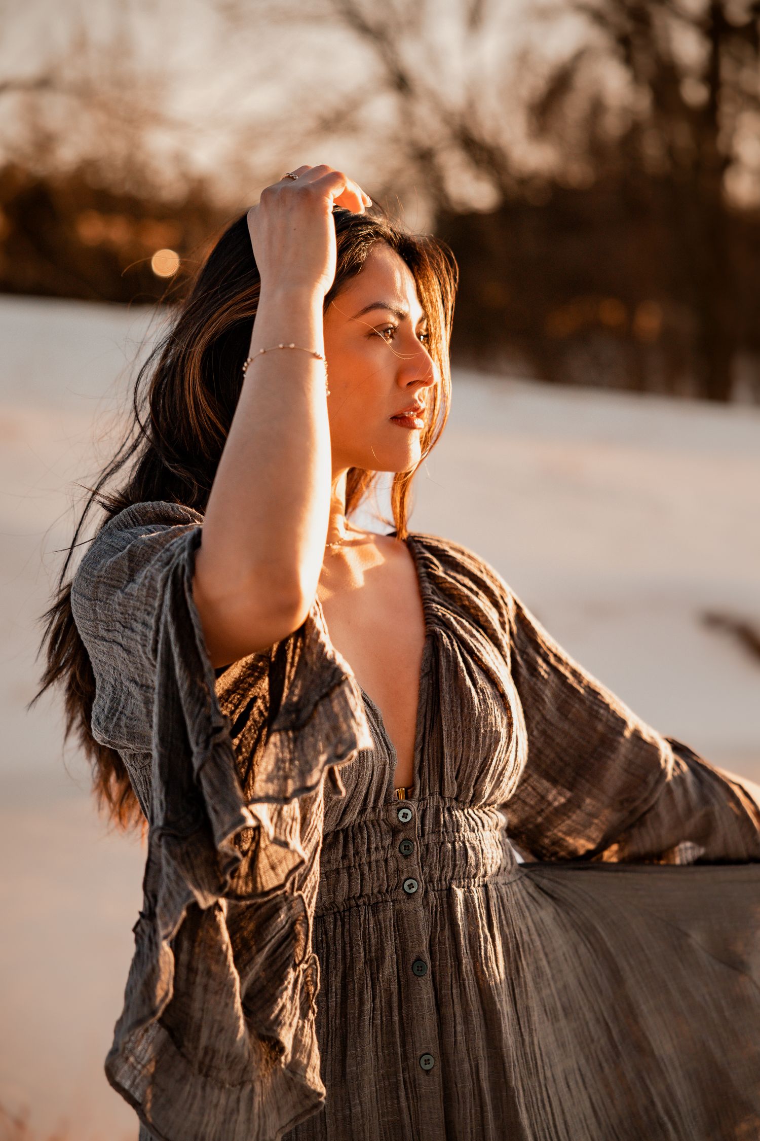 Woman in boho gray dress poses outdoors at golden hour, hand raised to head, with snowy landscape background.