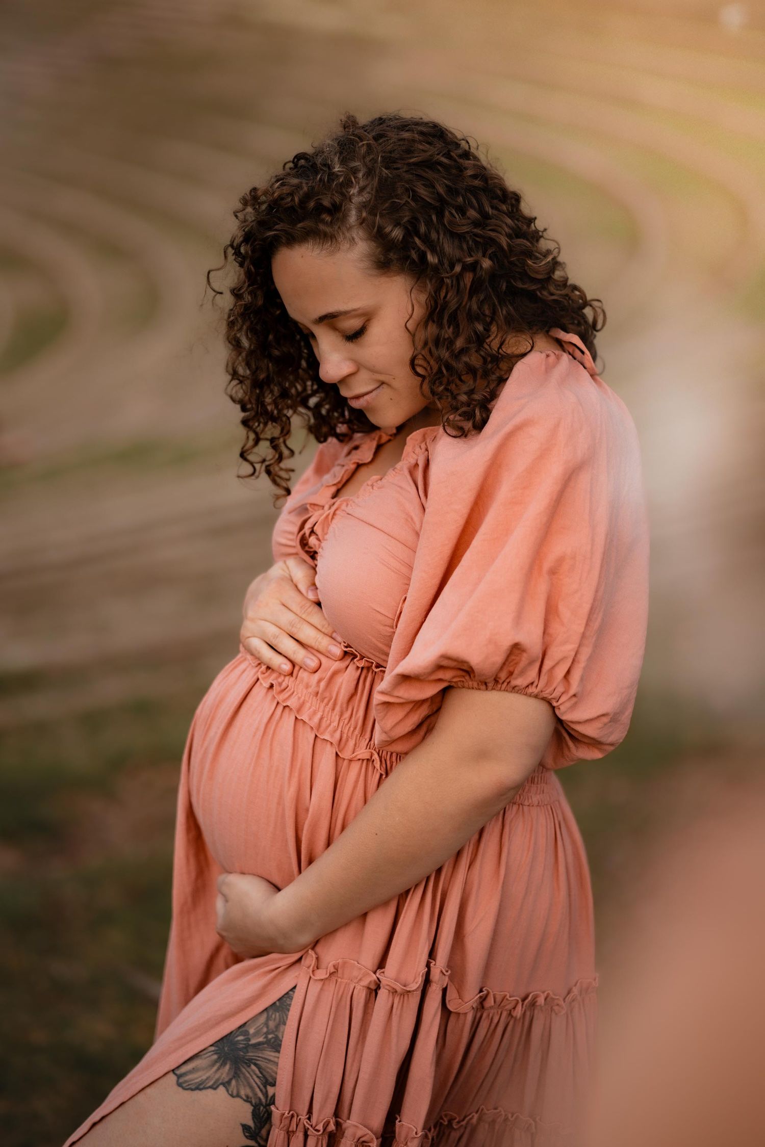 Pregnant woman in pink dress cradling baby bump outdoors at golden hour with curly hair and floral tattoo.