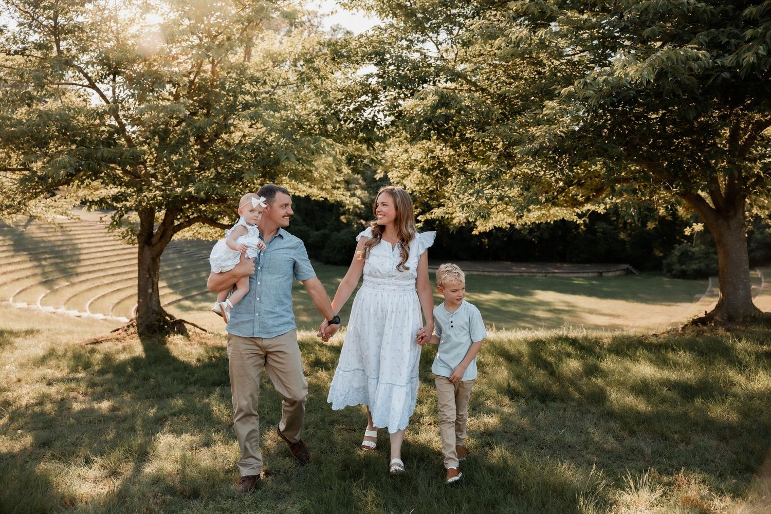 Family of four walking outdoors in a sunny park, parents holding hands with two young children among lush green trees.