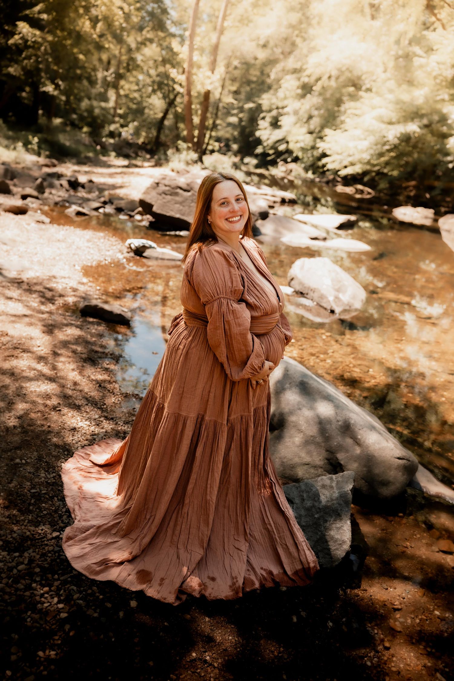 Smiling pregnant woman in flowing dusty rose maxi dress poses by rocky creek surrounded by lush green trees.