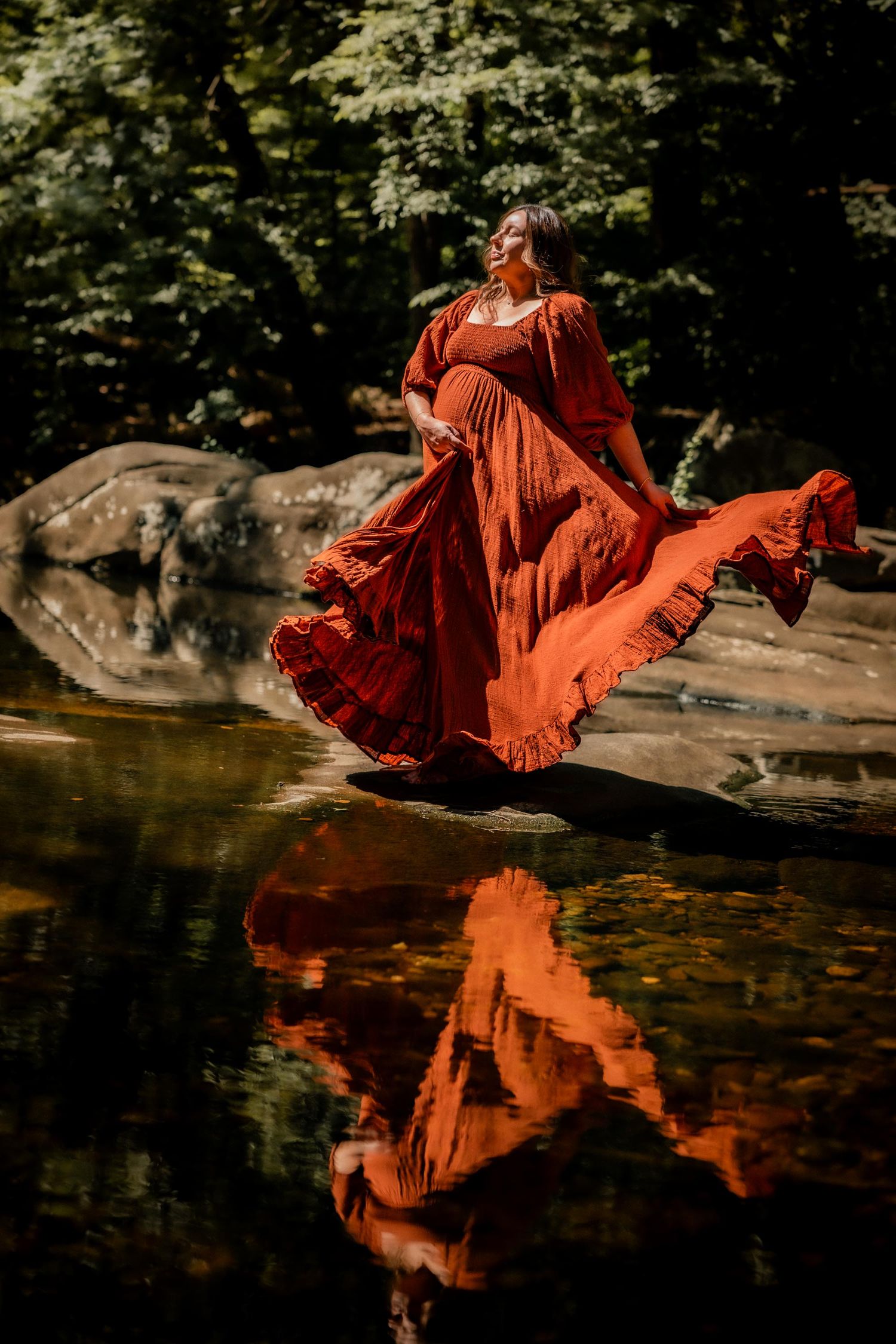 Pregnant woman in flowing red dress standing on rocks by a reflective forest stream, dress billowing dramatically.