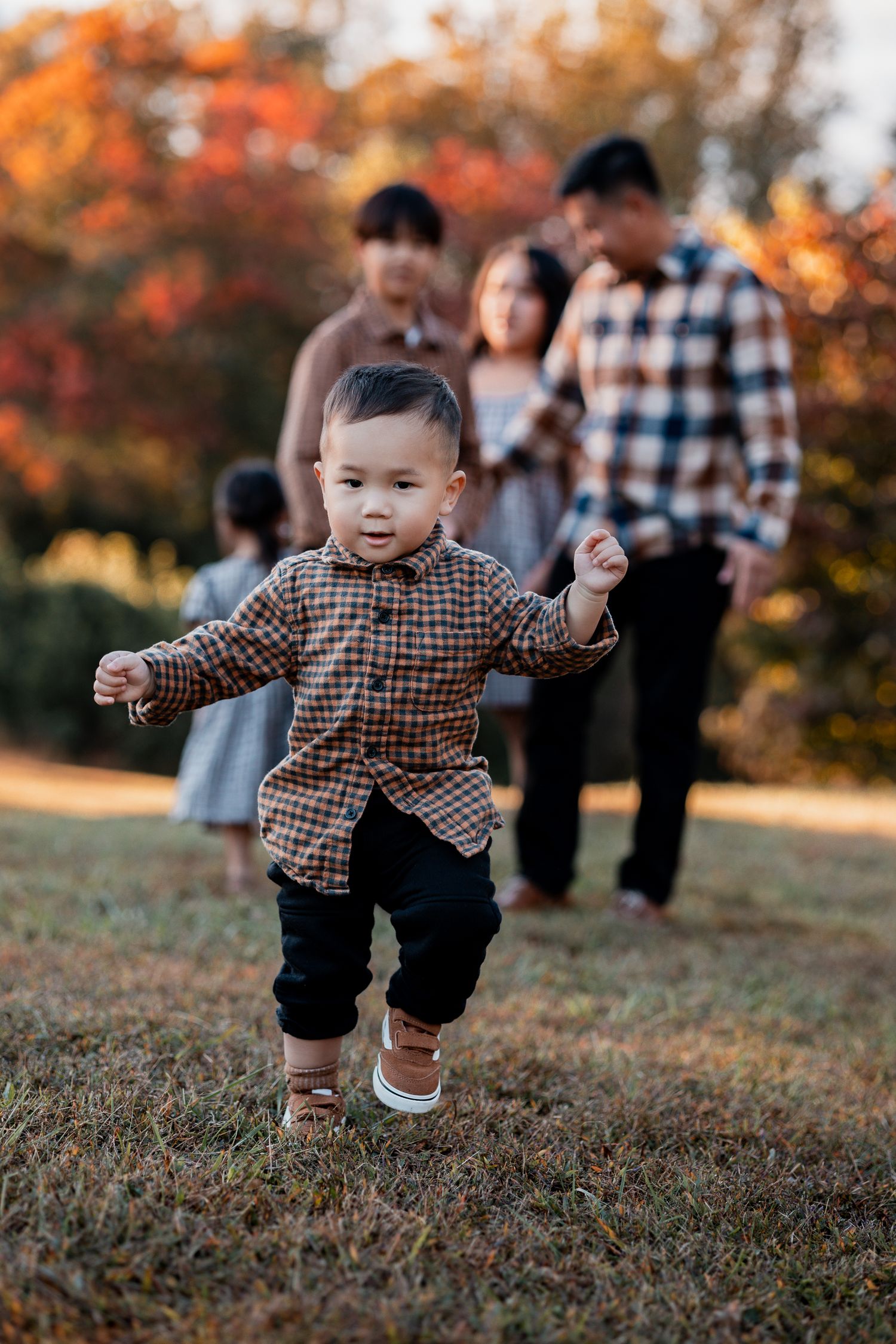 A toddler in a plaid shirt runs joyfully through autumn grass with colorful fall foliage in the background.