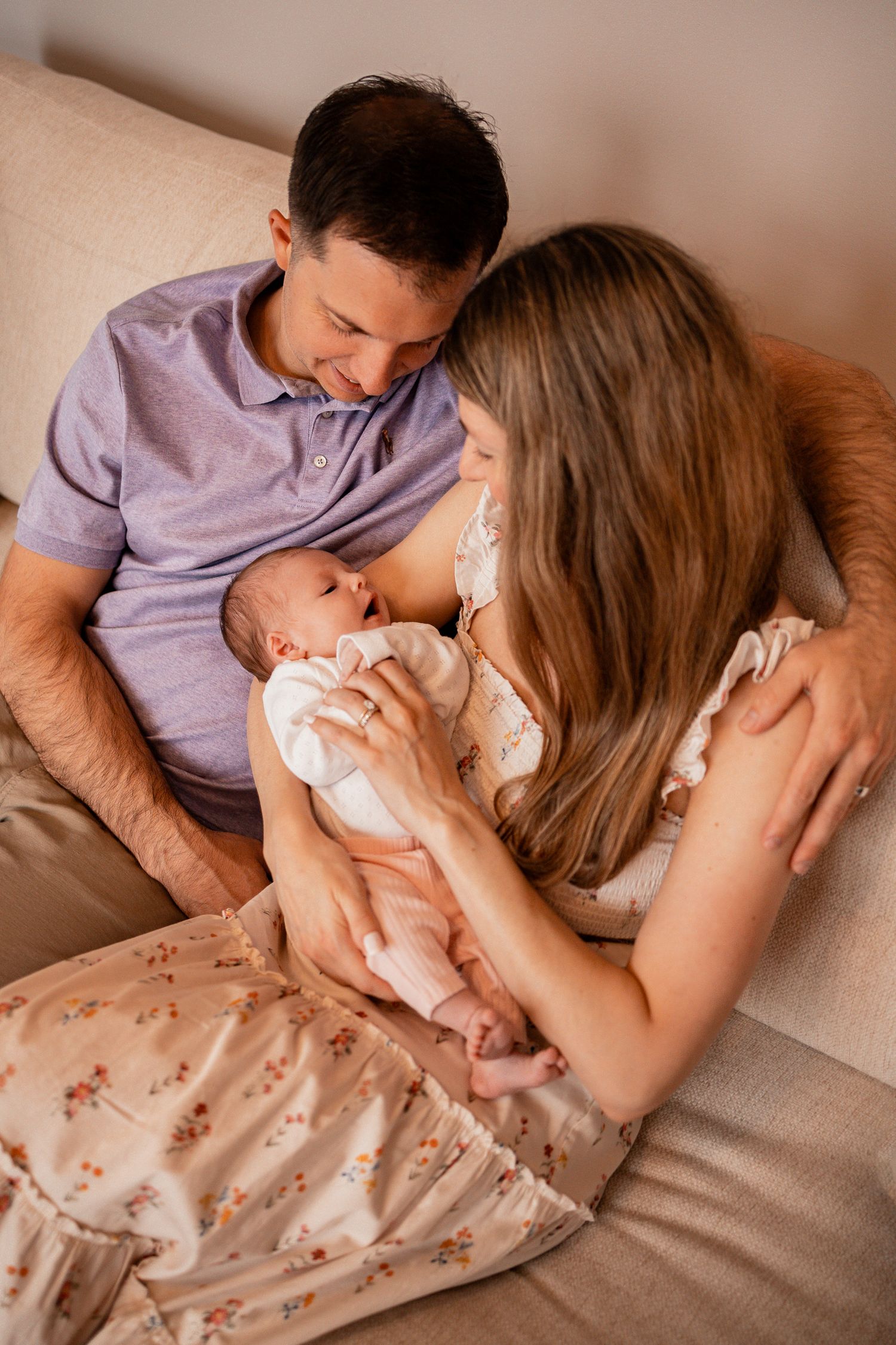 Intimate lifestyle photos of parents bonding with their newborn on a beige couch in warm lighting.