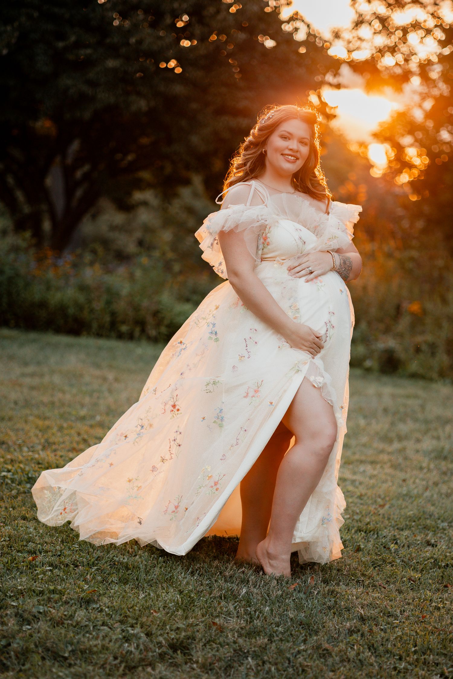 A pregnant woman in a flowing white lace dress poses in a garden at sunset with golden backlighting.