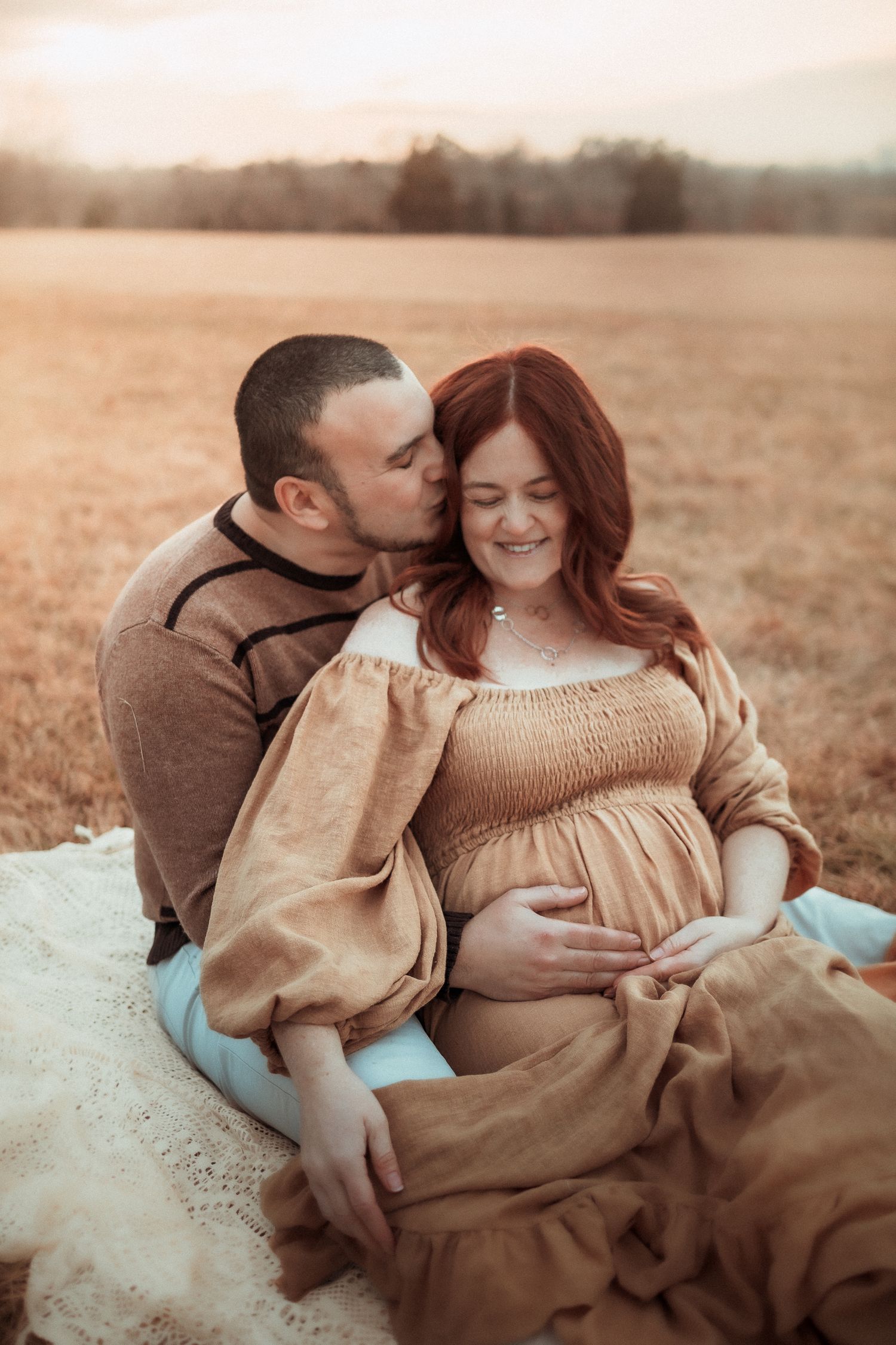 A tender maternity photoshoot in a field with warm tones, featuring a couple in casual earth-toned clothing.