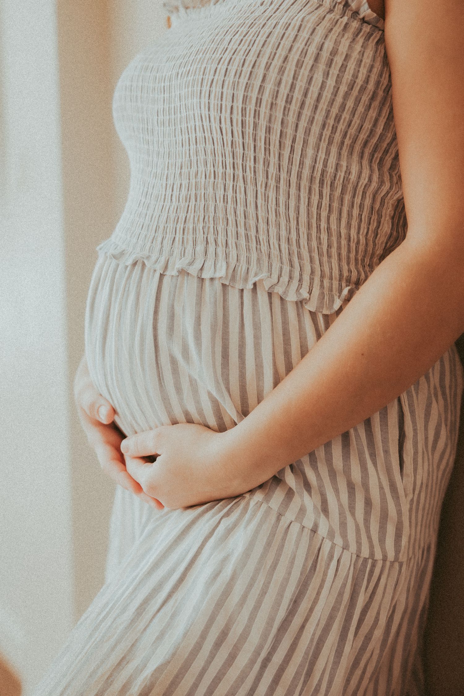 A pregnant woman in a soft white ribbed dress gently cradles her baby bump in natural window light.
