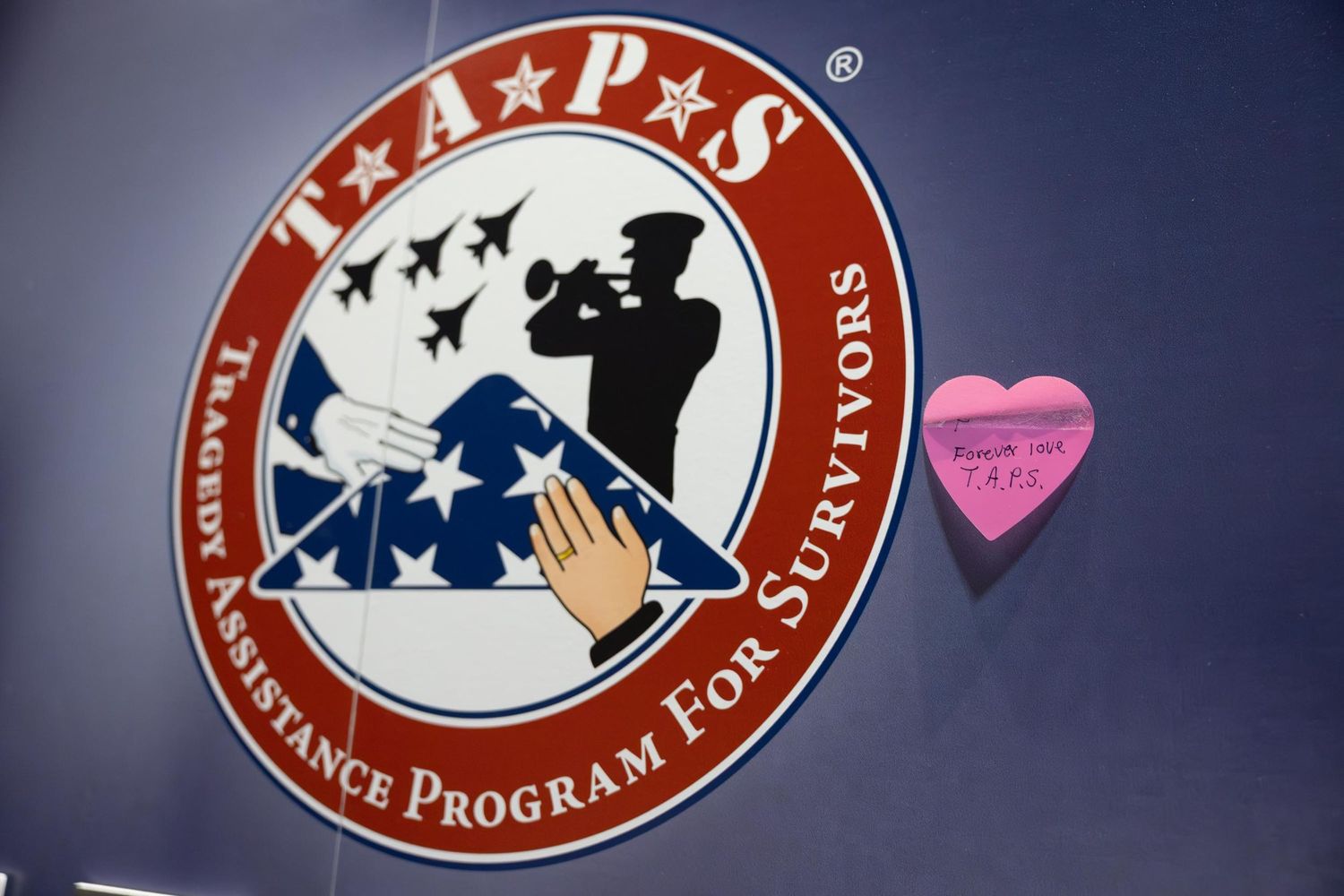 TAPS Tragedy Assistance Program for Survivors logo on a blue wall with a pink heart sticky note beside it.