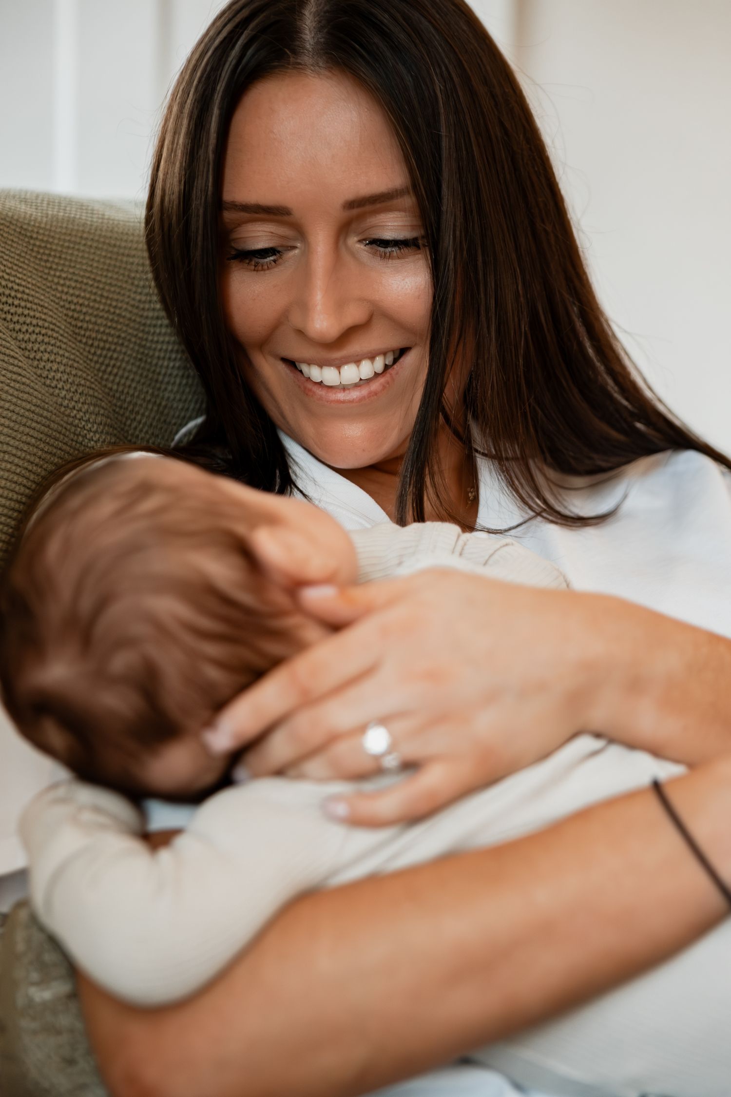 Mother wearing white sweater lovingly holds and smiles at sleeping baby while sitting in cozy armchair.