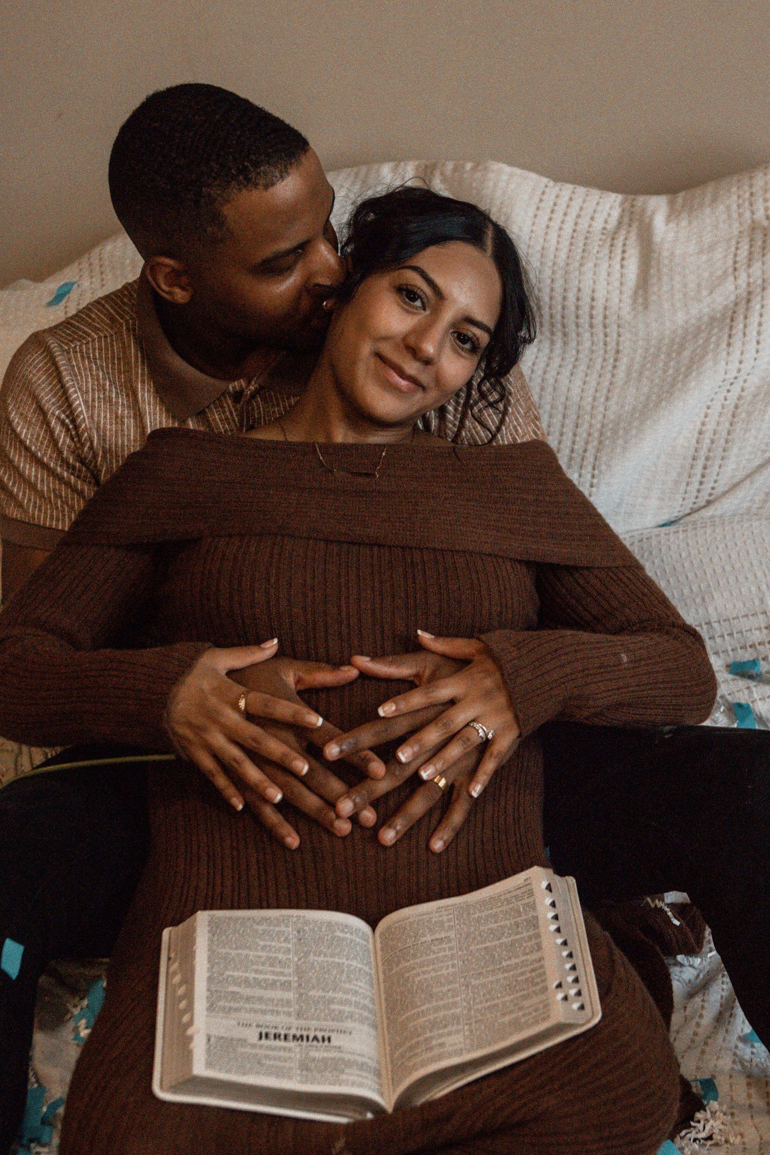A cozy couple sits on a couch embracing while reading a Bible together in warm brown sweaters.