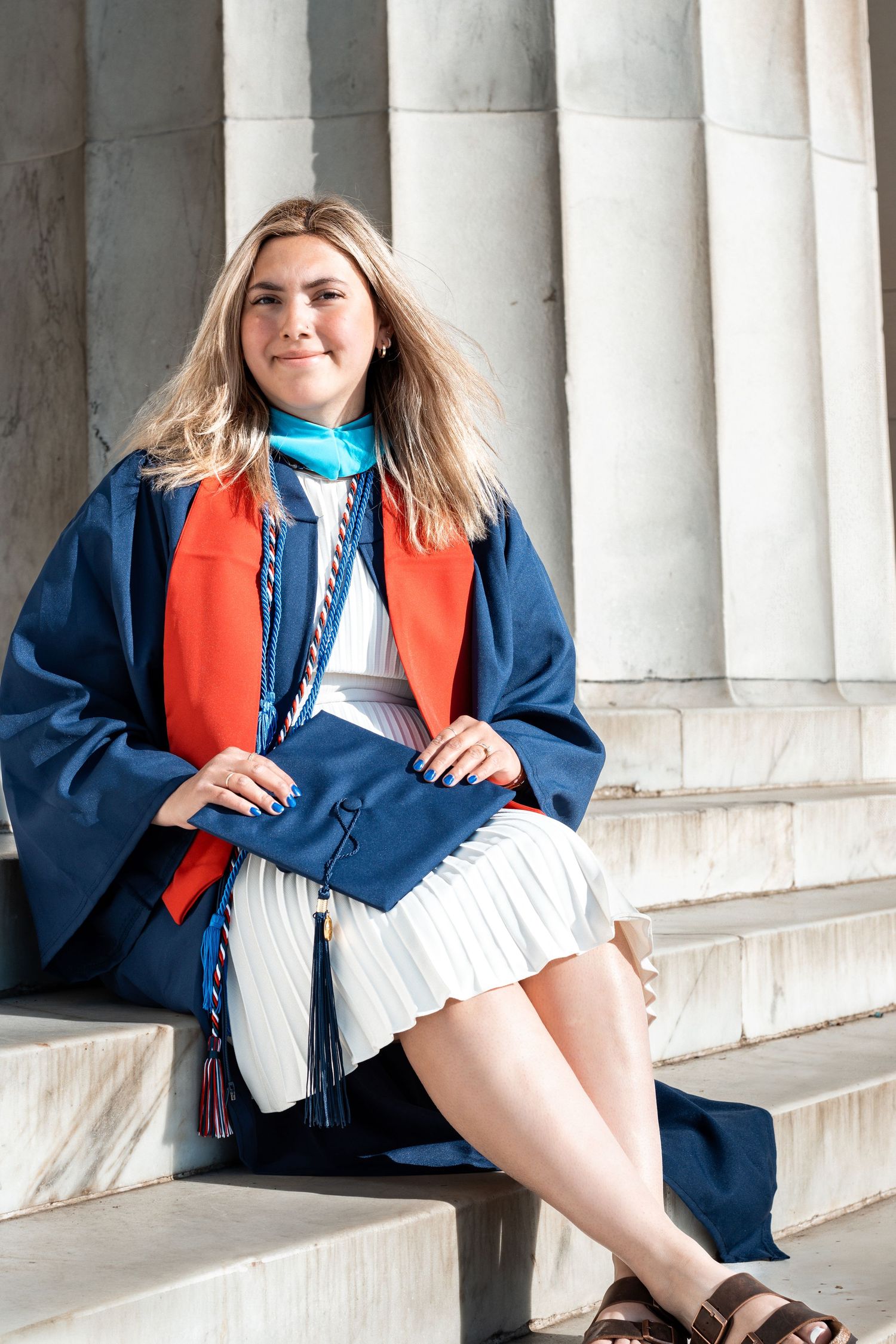 Graduate in cap and gown sits on white stone steps in classic architectural setting.