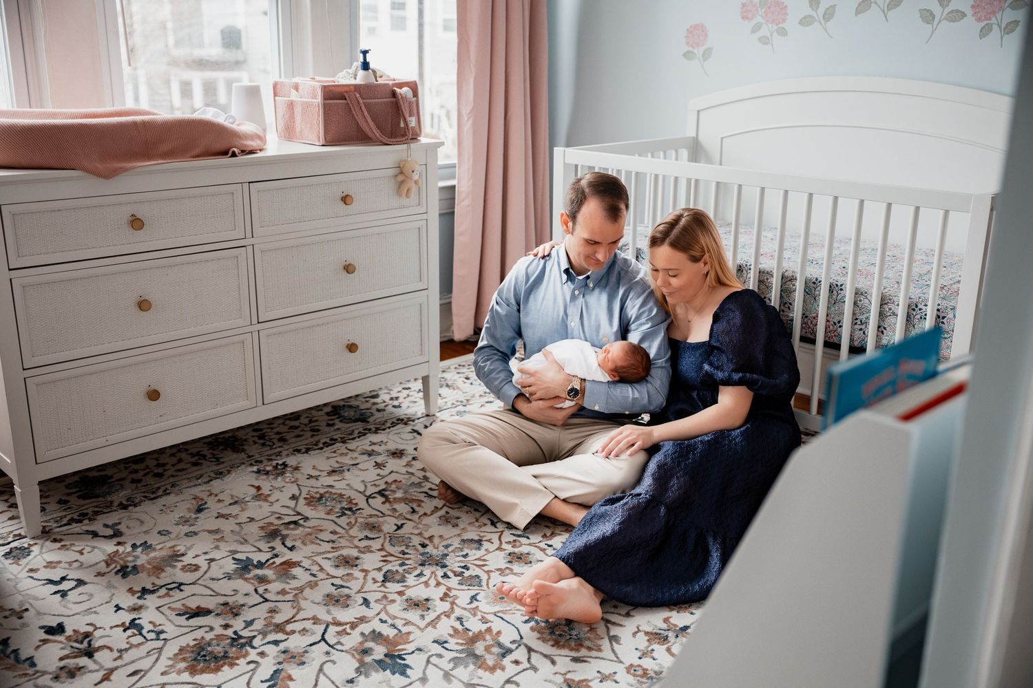 Parents sitting on nursery floor with newborn baby, white crib and dresser in background.