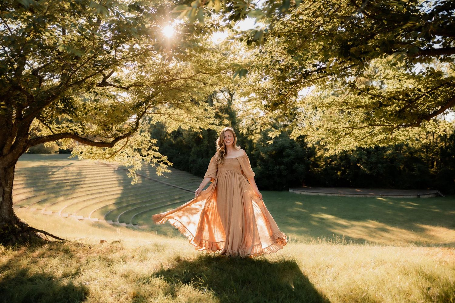 Woman in flowing peach gown twirling under sunlit trees in a lush green park setting.