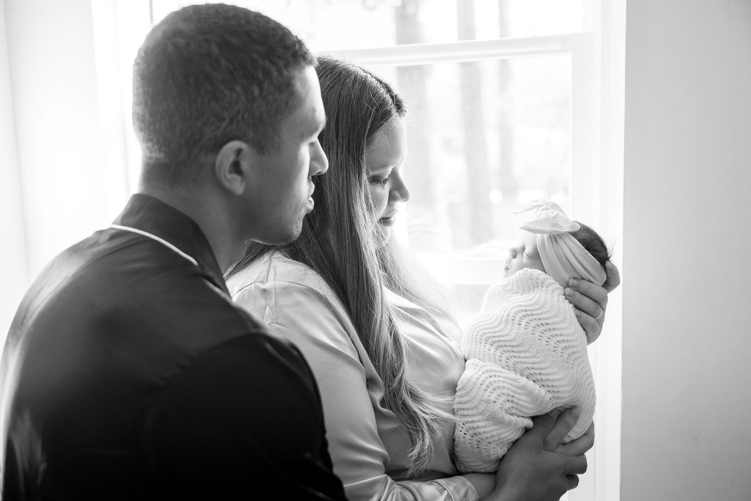 Black and white photo of new parents lovingly gazing at their swaddled newborn baby by a bright window.
