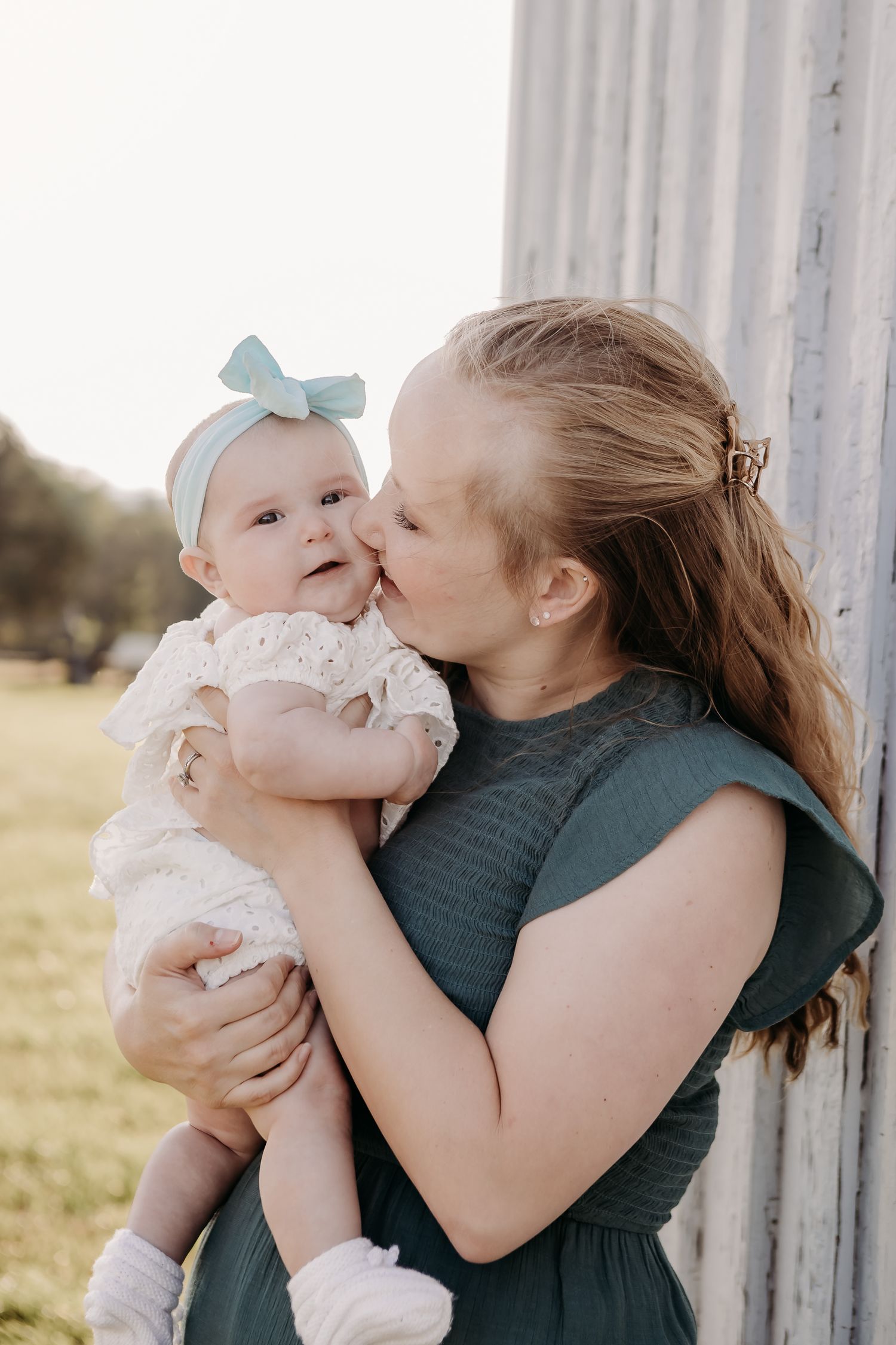 A tender moment captured outdoors as a mother in a green dress kisses her baby wearing a white dress and blue bow.