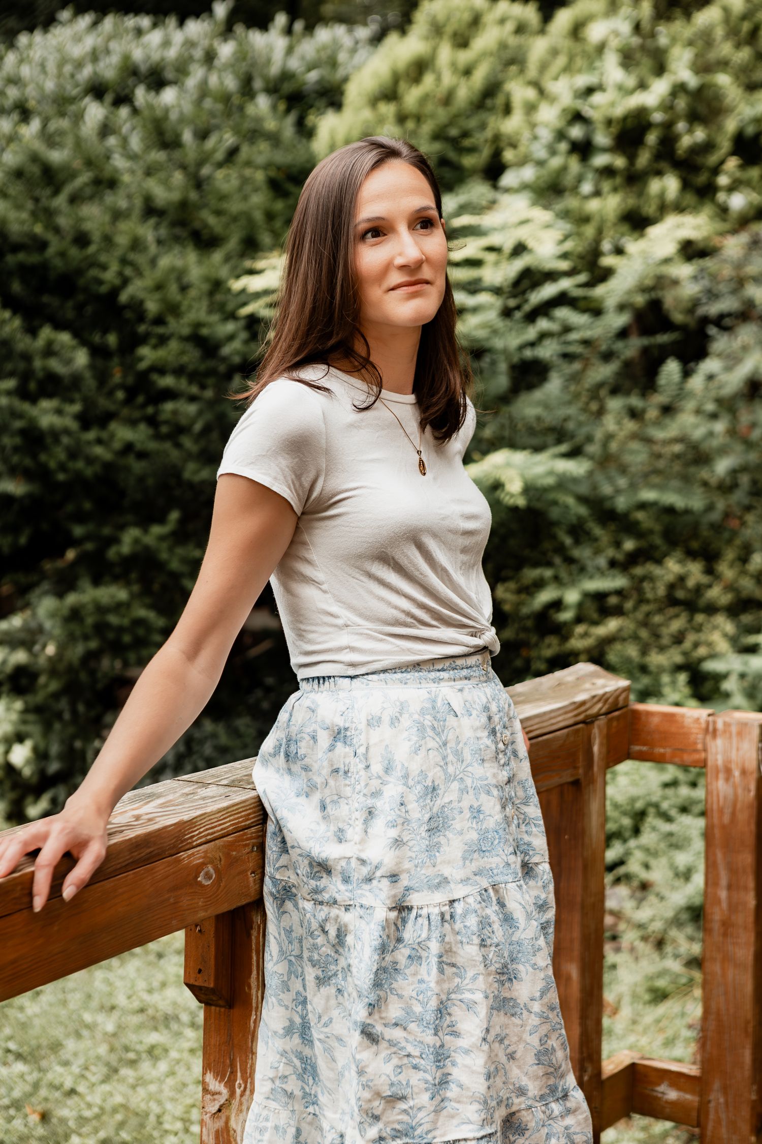 A person in a floral skirt and cream colored top stands against a wooden railing surrounded by lush greenery.