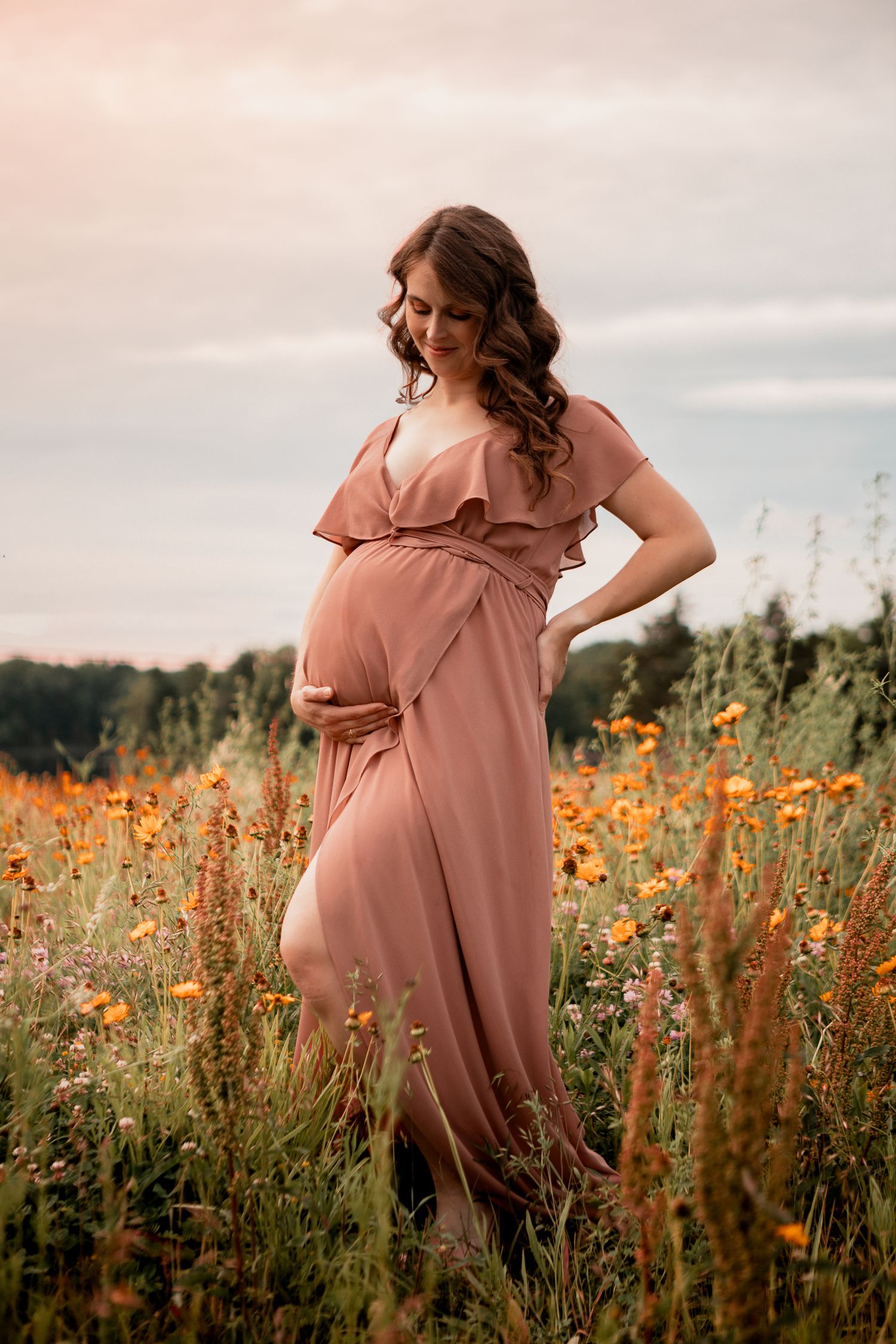 Beautiful maternity portrait in a dusty pink dress standing among wildflowers and orange cosmos at sunset.