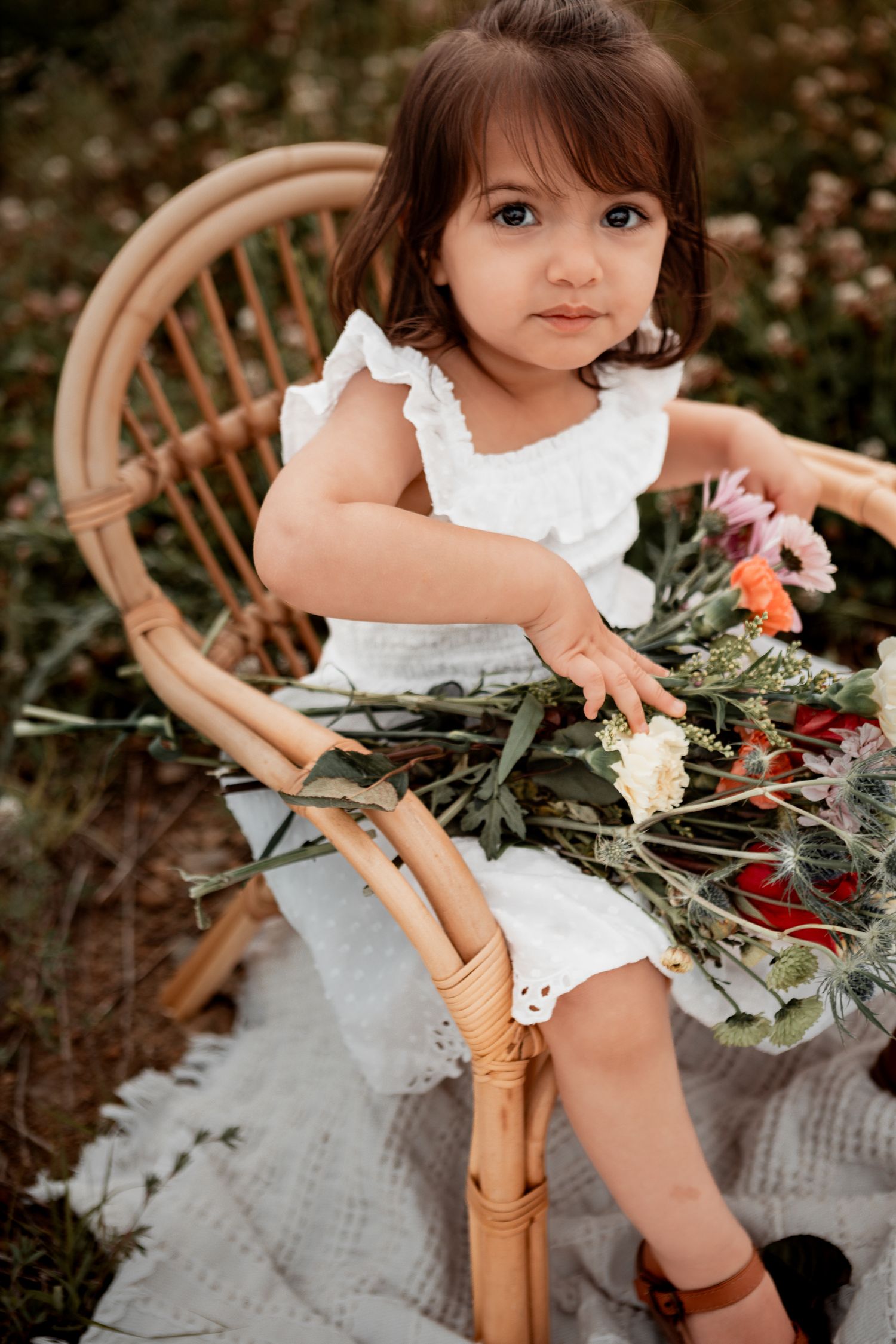 A small child in white ruffled dress holds flowers while sitting in a wooden chair against a dark backdrop.