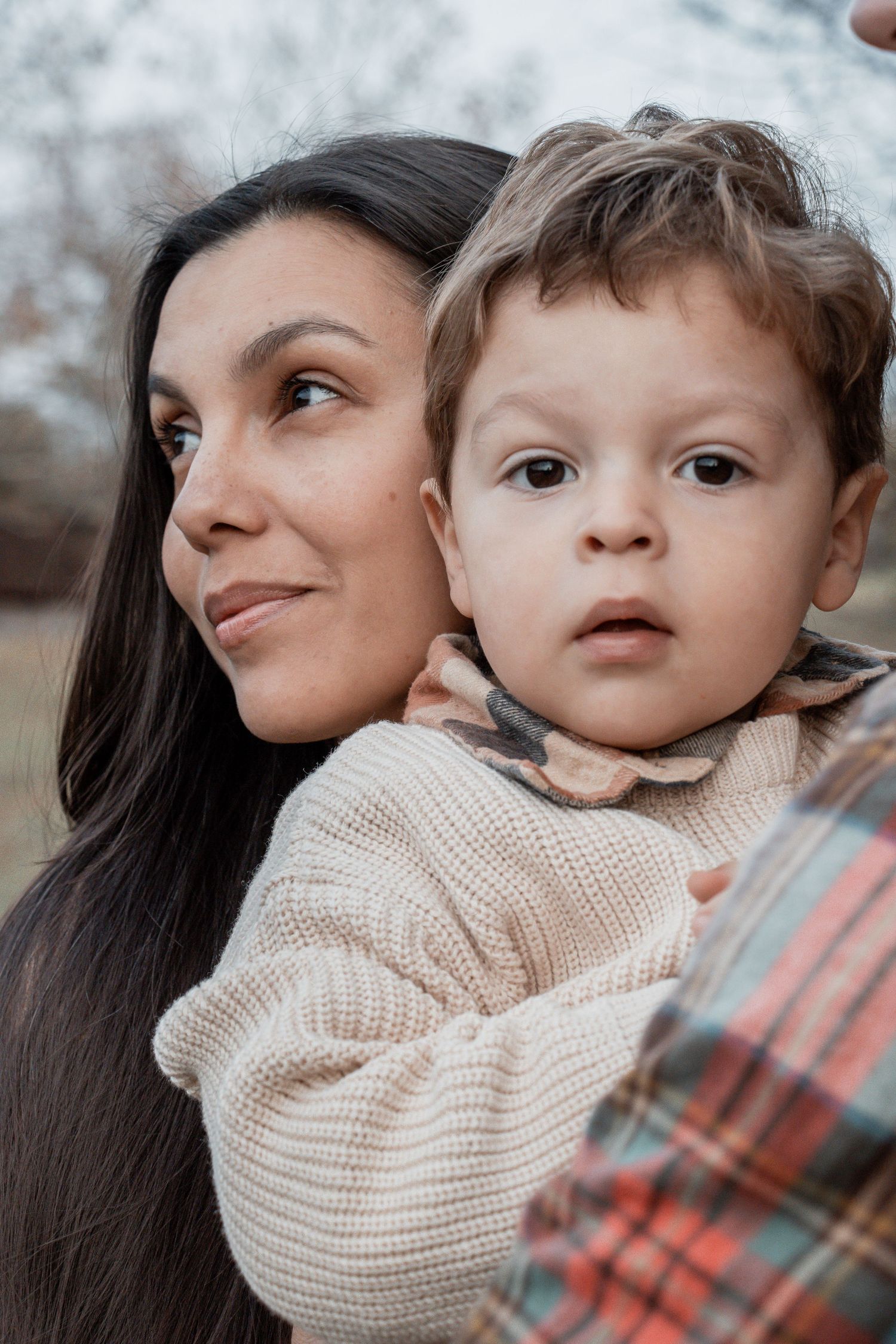 A person with long dark hair holds a young child wearing a cream knit sweater on a winter day.