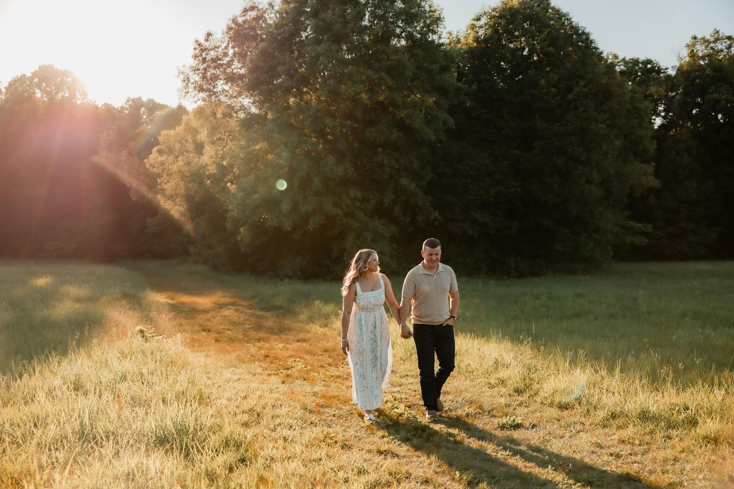 Couple holding hands walking through a sunlit meadow with golden hour light and lush green trees in background.