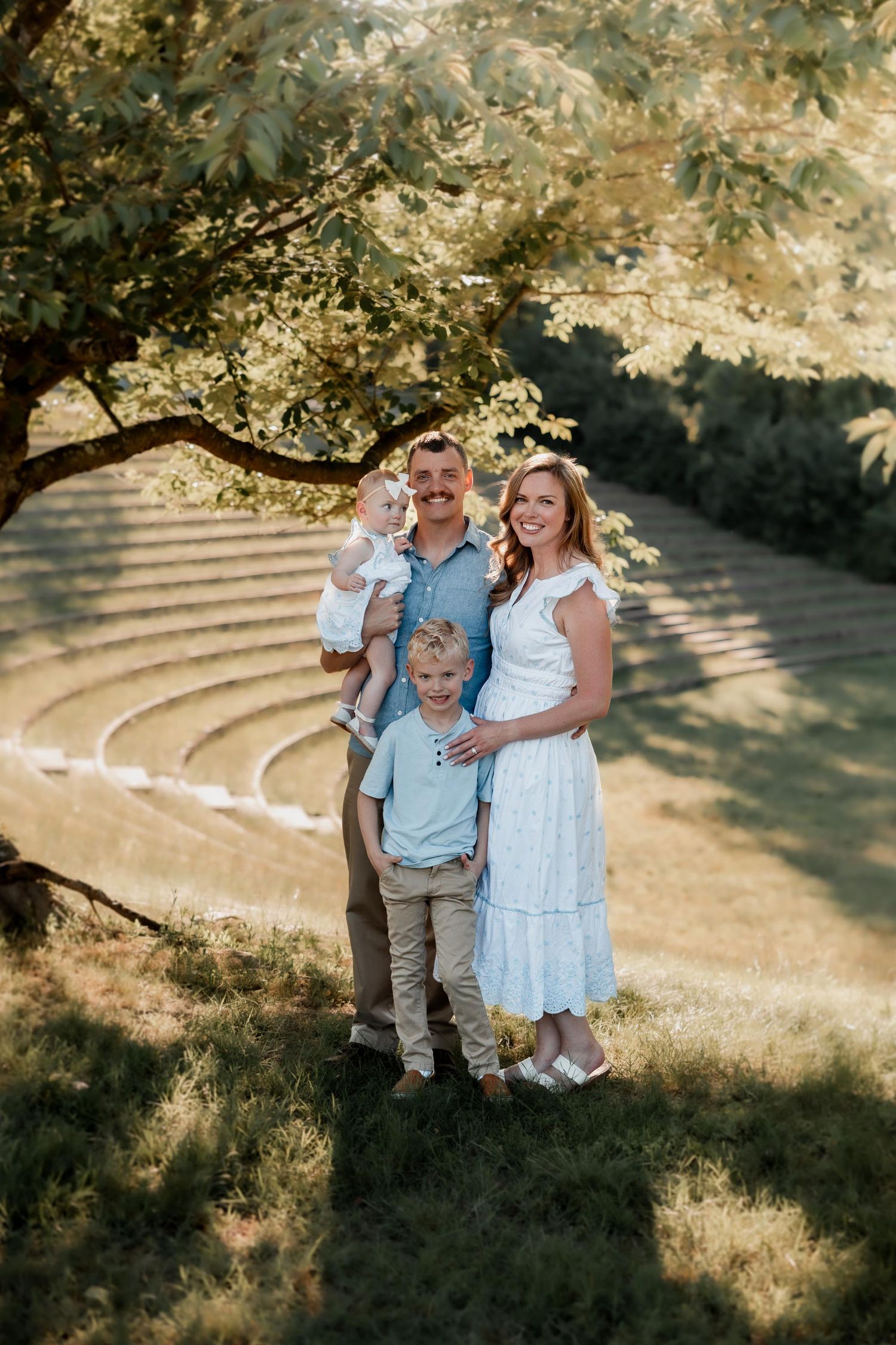 Happy family of four posing outdoors under a tree in a sunny park, parents and two young children smiling.