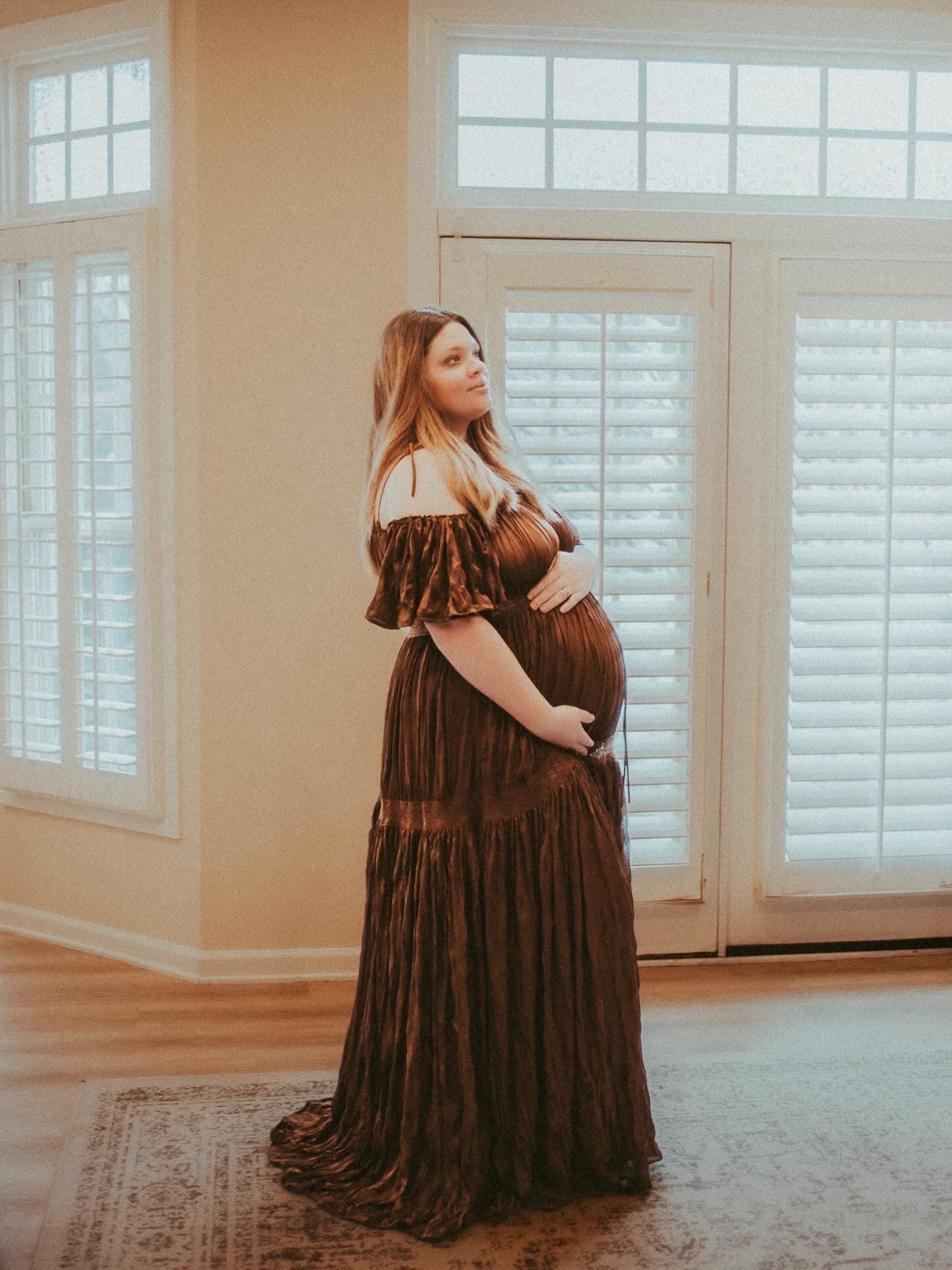 A person in a flowing brown dress poses gracefully against white shuttered windows in a bright interior space.