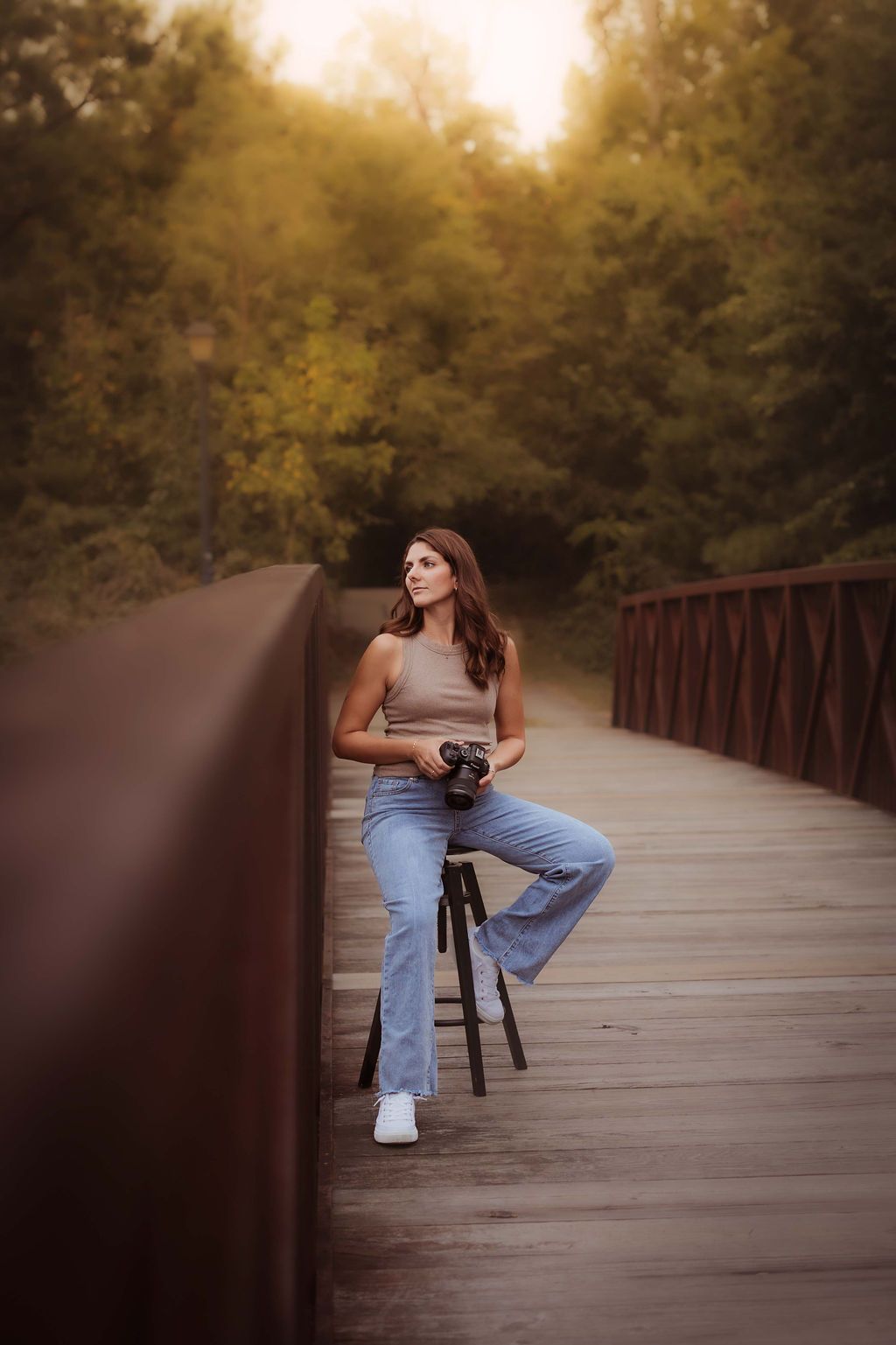 Woman photographer sitting on bridge with camera and tripod, surrounded by lush green trees in warm golden light.