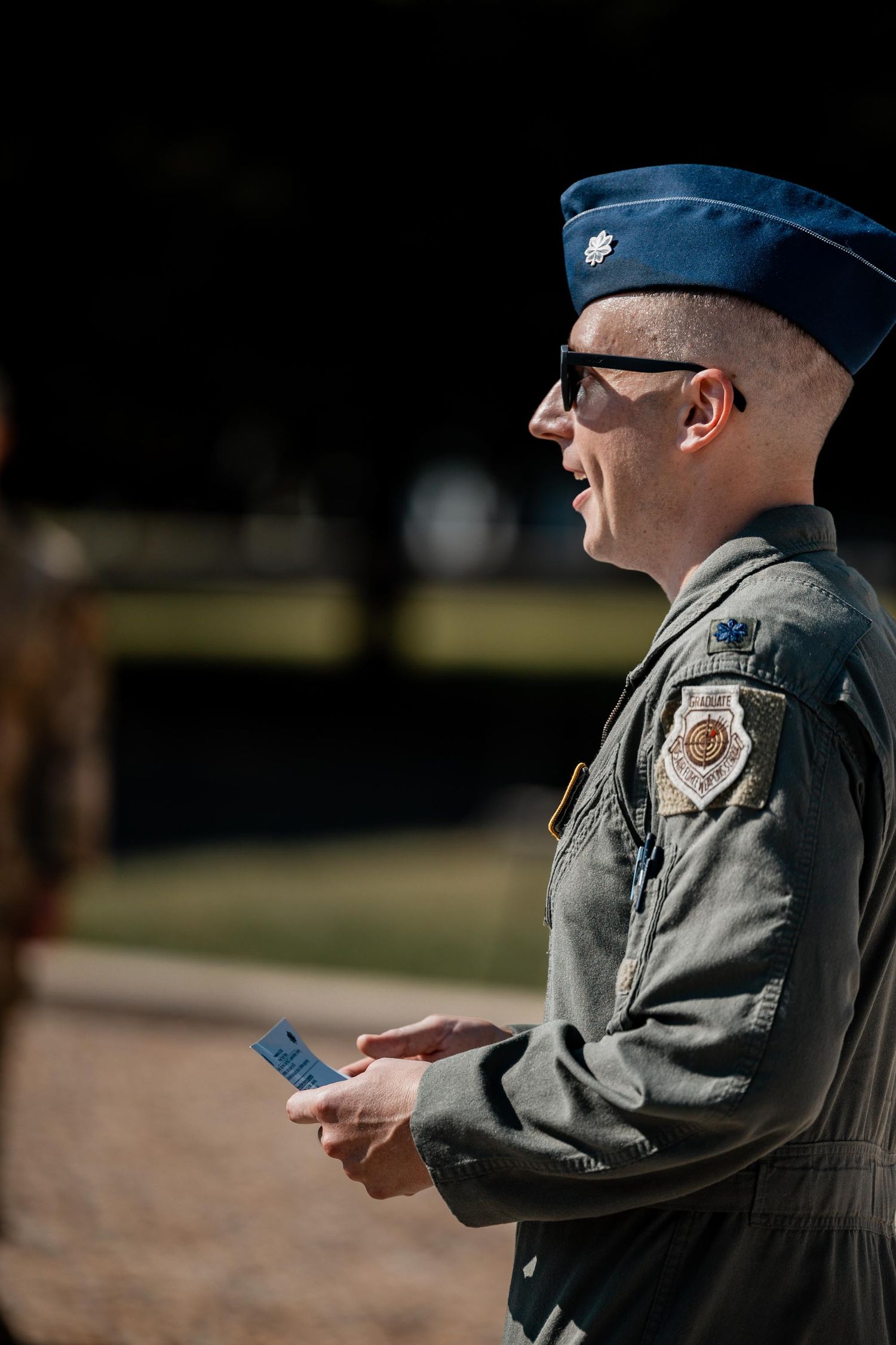 Military officer in flight suit and blue cap holding a card outdoors in sunlight.