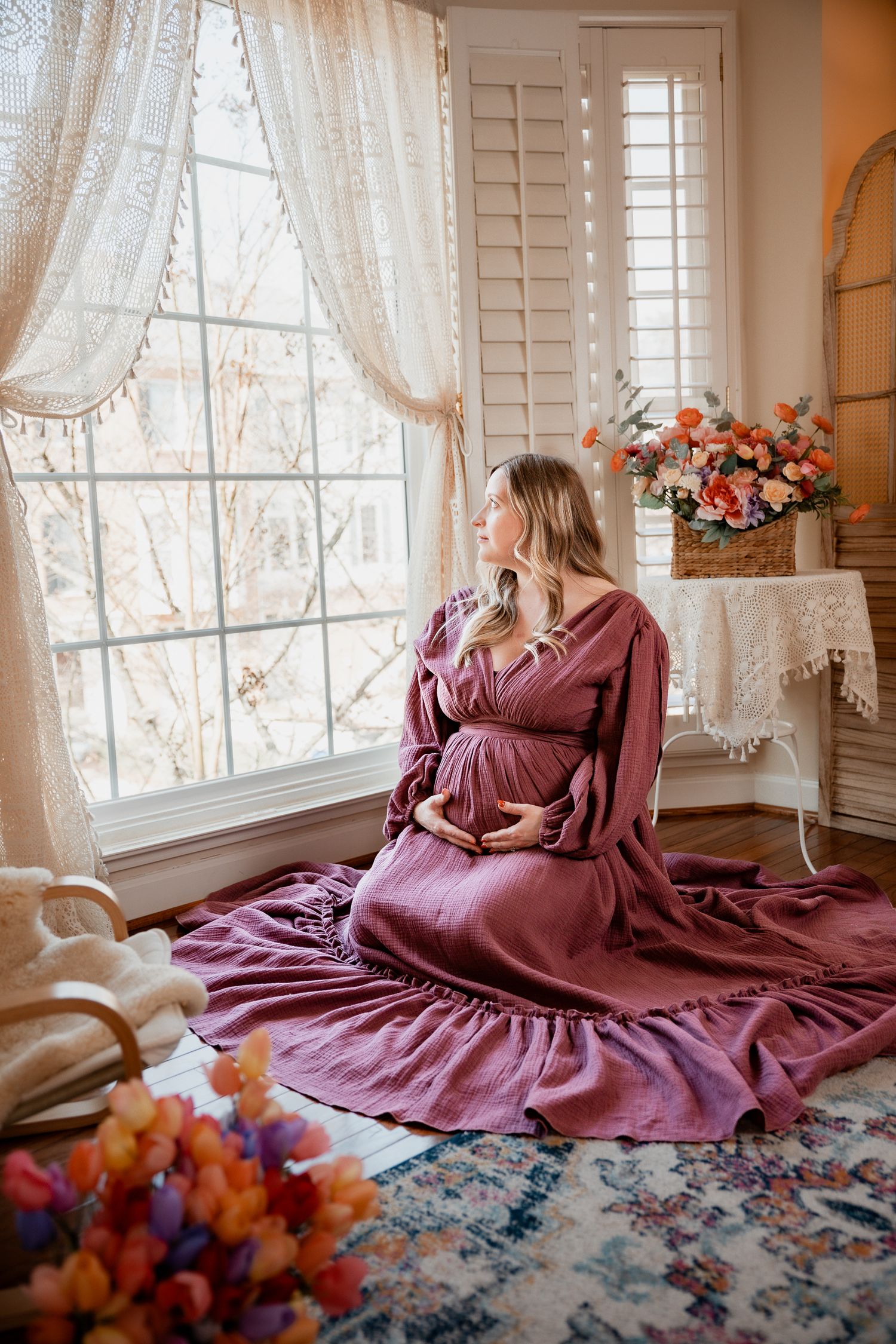 Pregnant woman in mauve dress sitting on floor by large window, cradling baby bump in elegant home setting.