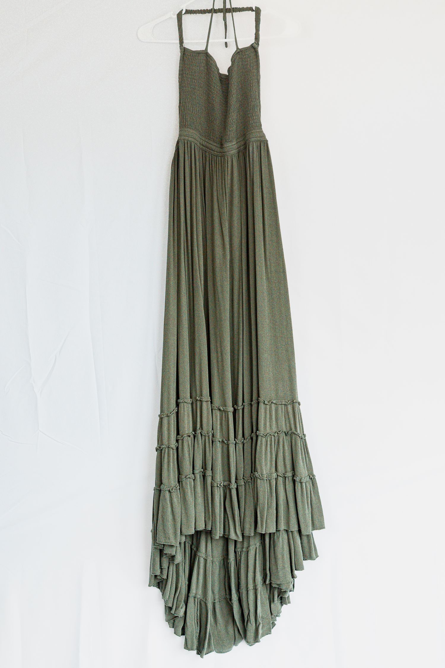 Olive green maxi dress with tiered ruffle hem and spaghetti straps hangs against white wall.