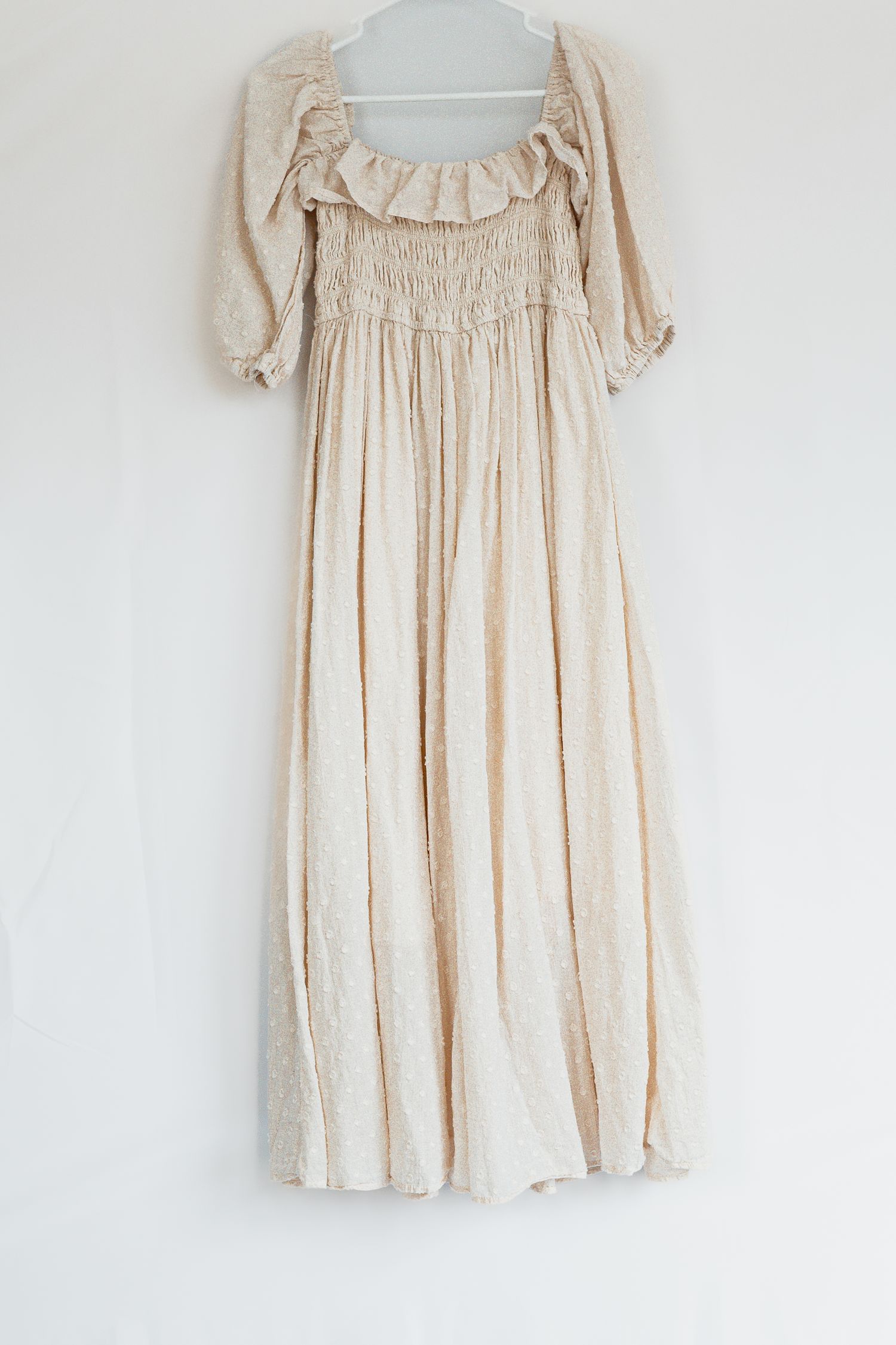 Romantic cream-colored smocked maxi dress with ruffled neckline and three-quarter length sleeves hanging on white wall.