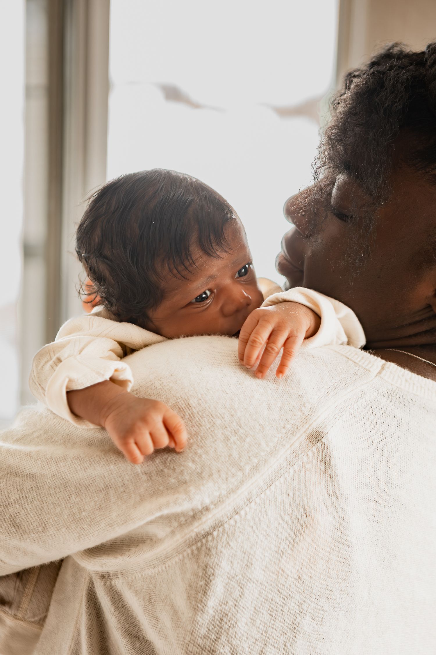 A tender parenting moment with a baby in a cream sweater peeking over a shoulder in soft window light.