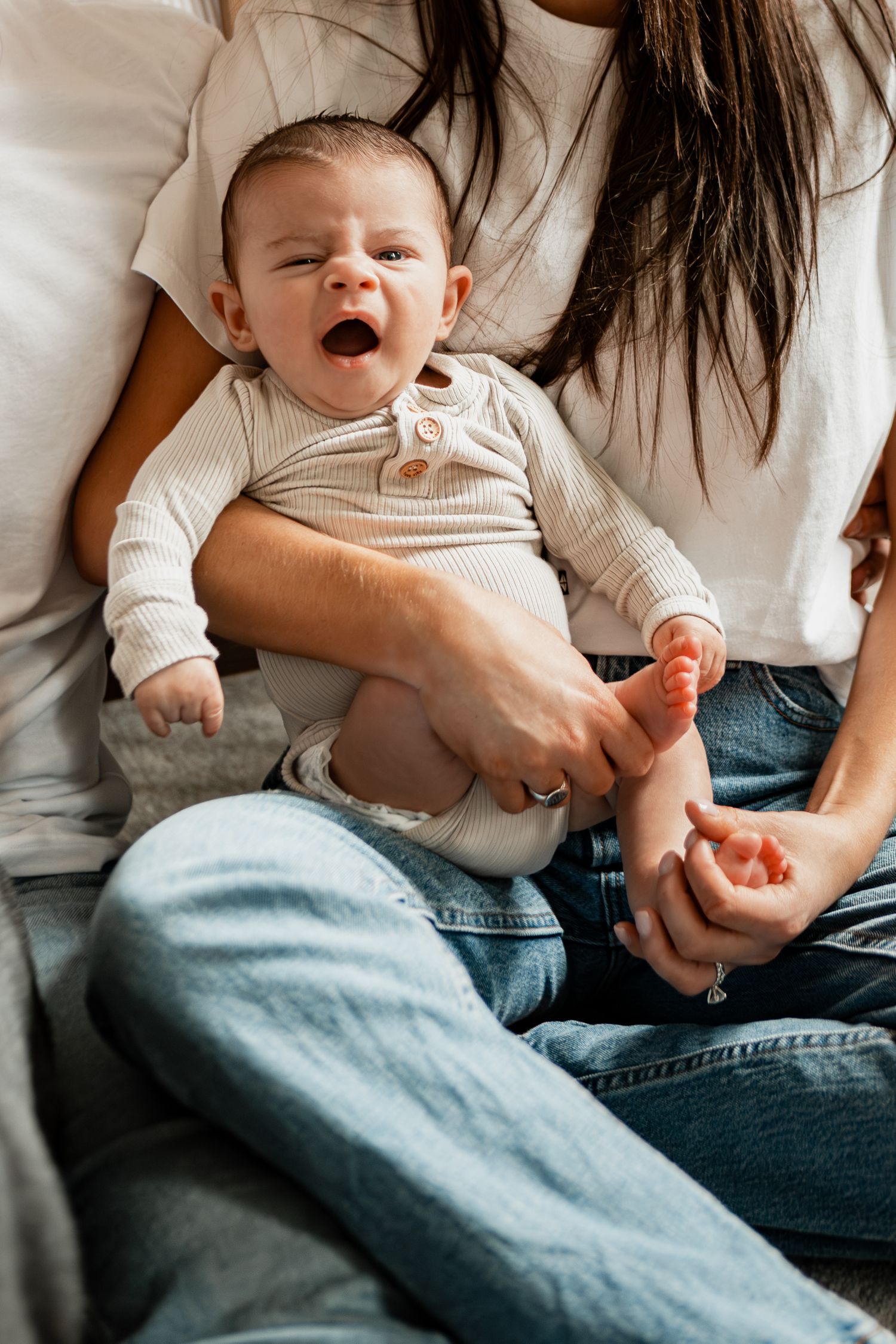 A yawning baby wearing a white onesie rests on someone's lap in casual denim jeans.