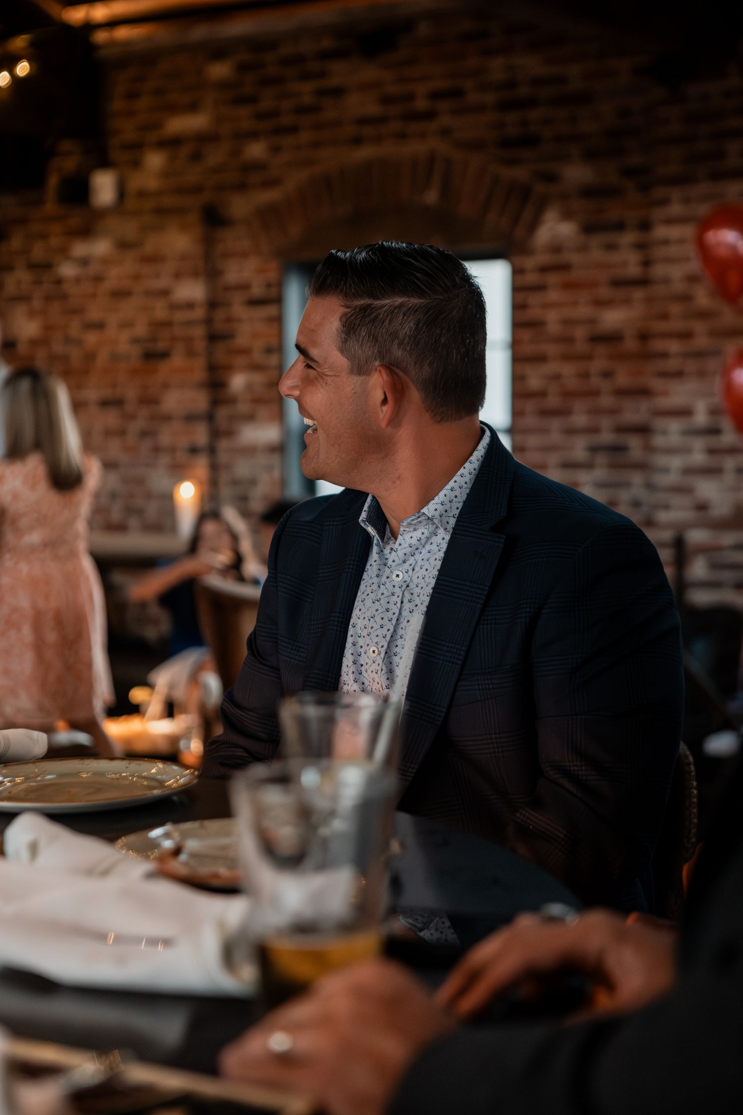 A guest in a navy blazer sits at a table during an evening event with brick wall ambiance.