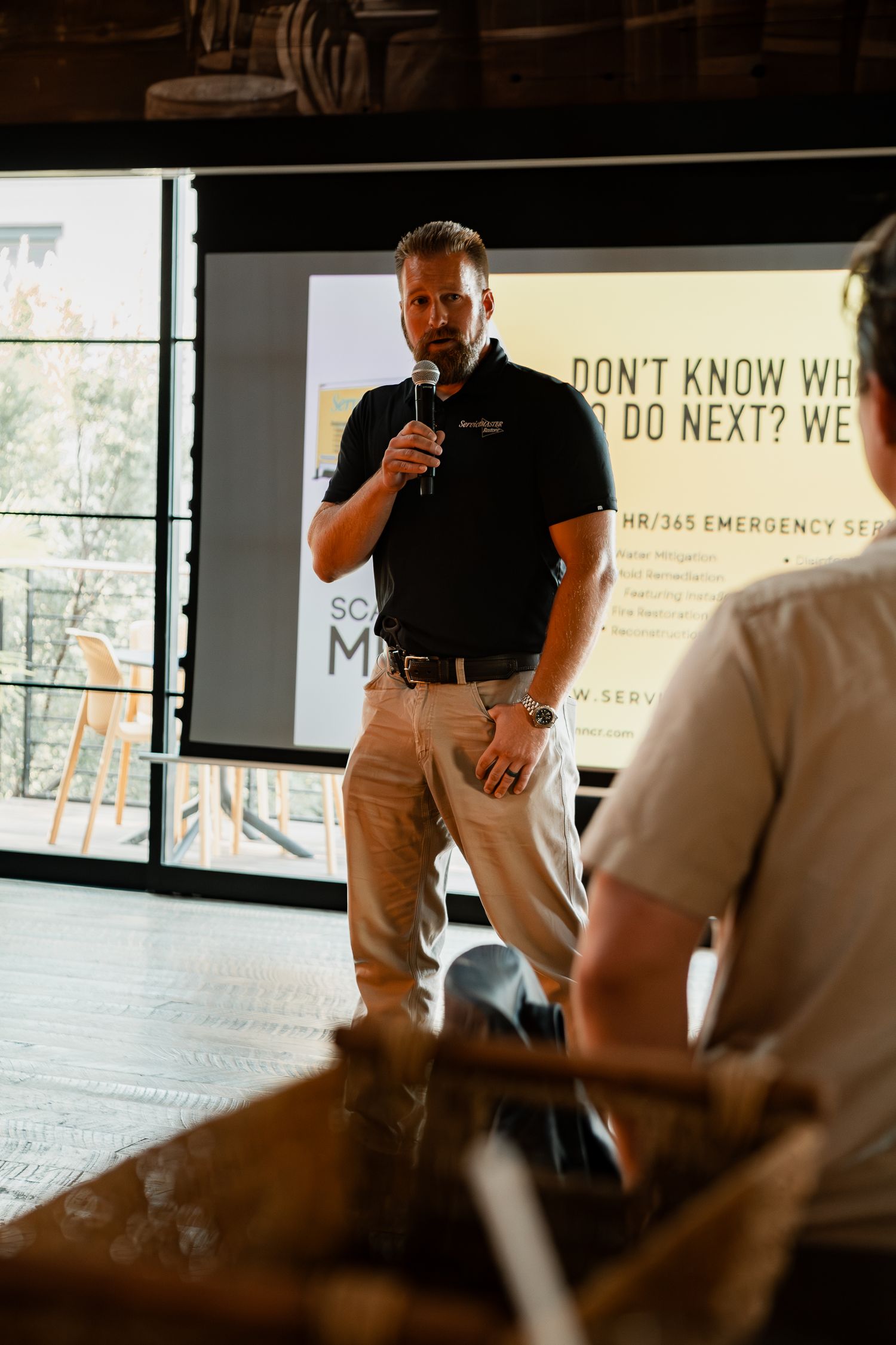 A presenter addresses an audience in a modern space with natural lighting.