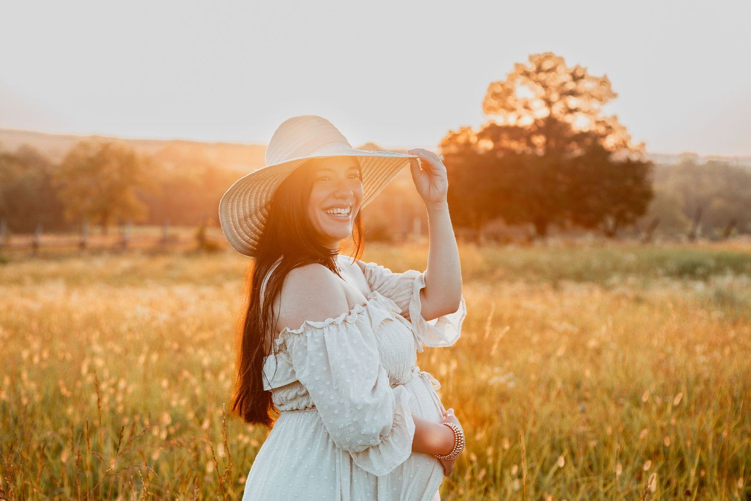 Someone in a white sundress and wide brim hat stands in a golden sunlit field at sunset.