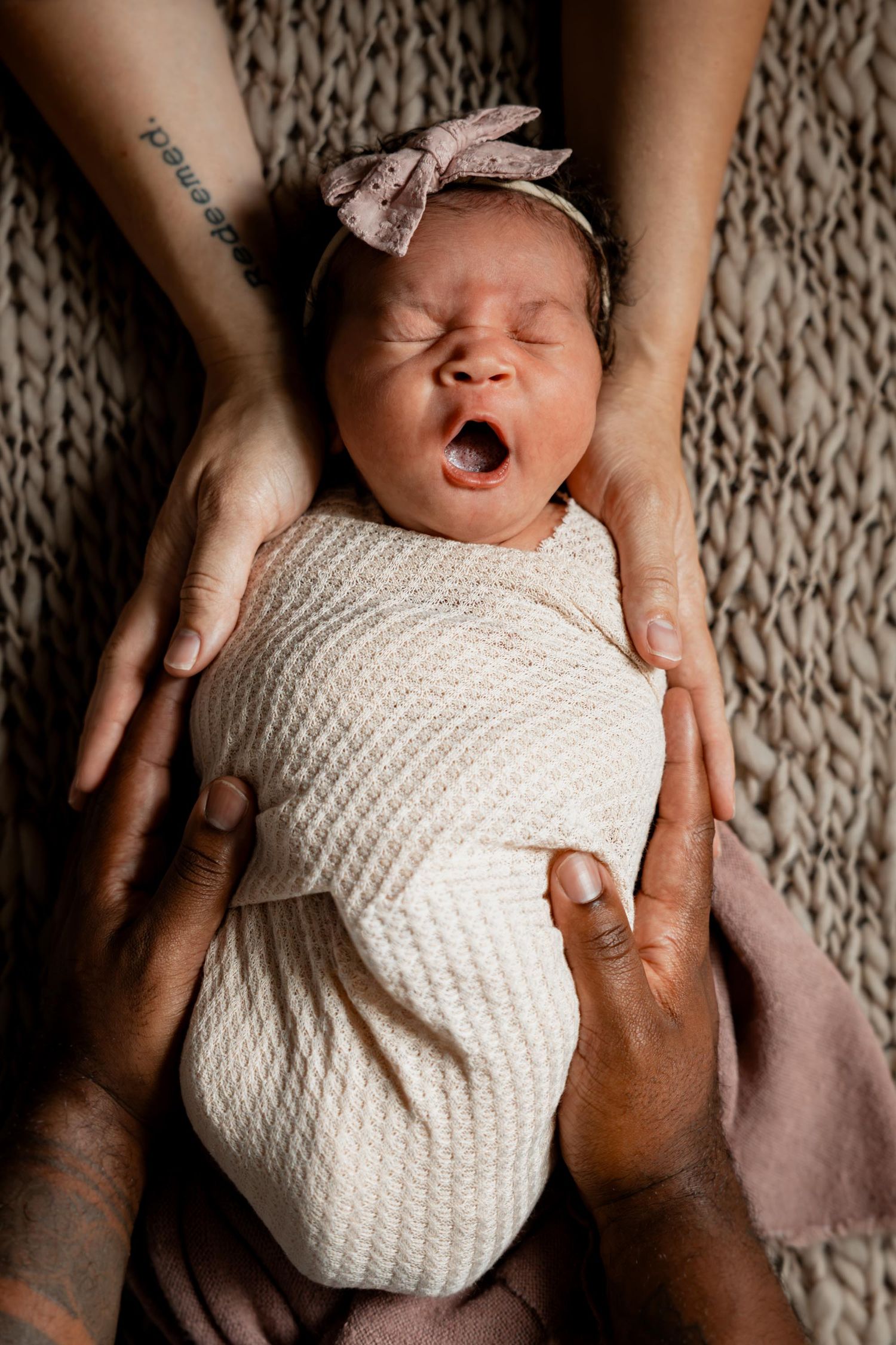 Newborn baby girl swaddled in white, wearing a pink bow headband, yawning while cradled by two pairs of loving hands.
