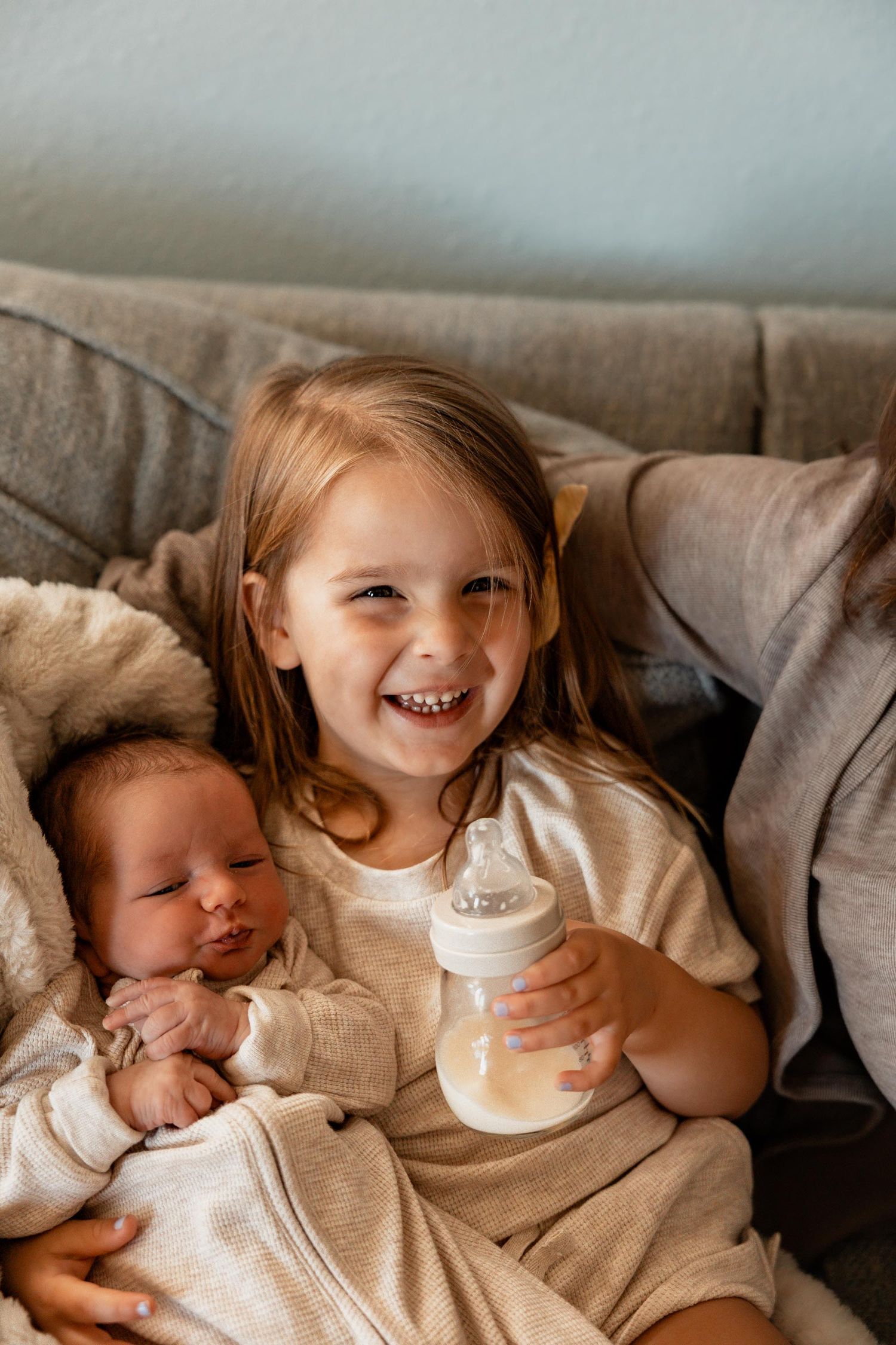 Young girl smiling on couch holding baby bottle next to sleeping newborn sibling wrapped in white blanket.