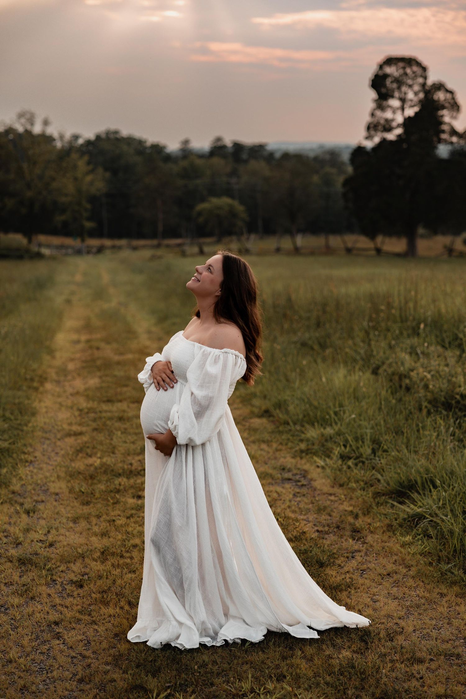 Glowing pregnant woman in flowing white off-shoulder gown standing on rural path at sunset, embracing her bump.