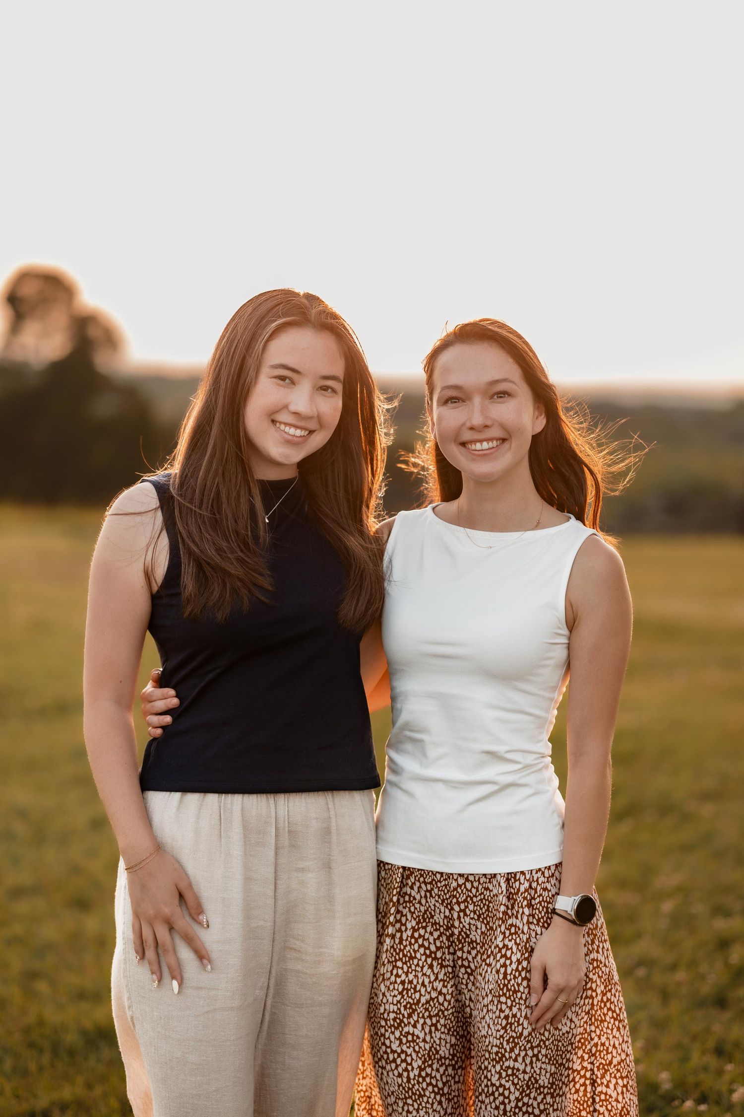 Two smiling young women pose together in a sunlit field during golden hour, one in black top and one in white top.