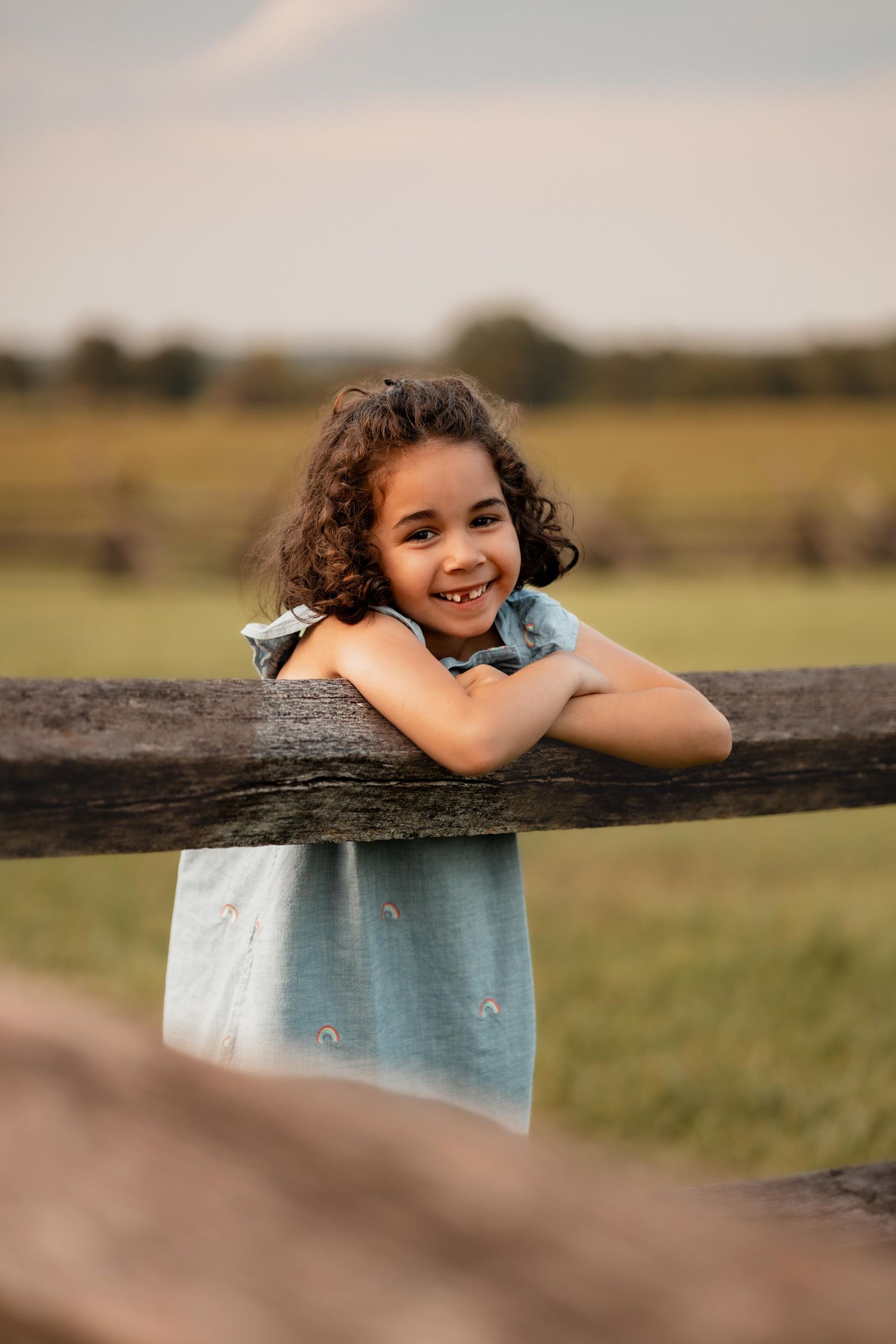 A smiling young girl with curly hair leans on a wooden fence rail in a sunny outdoor field.